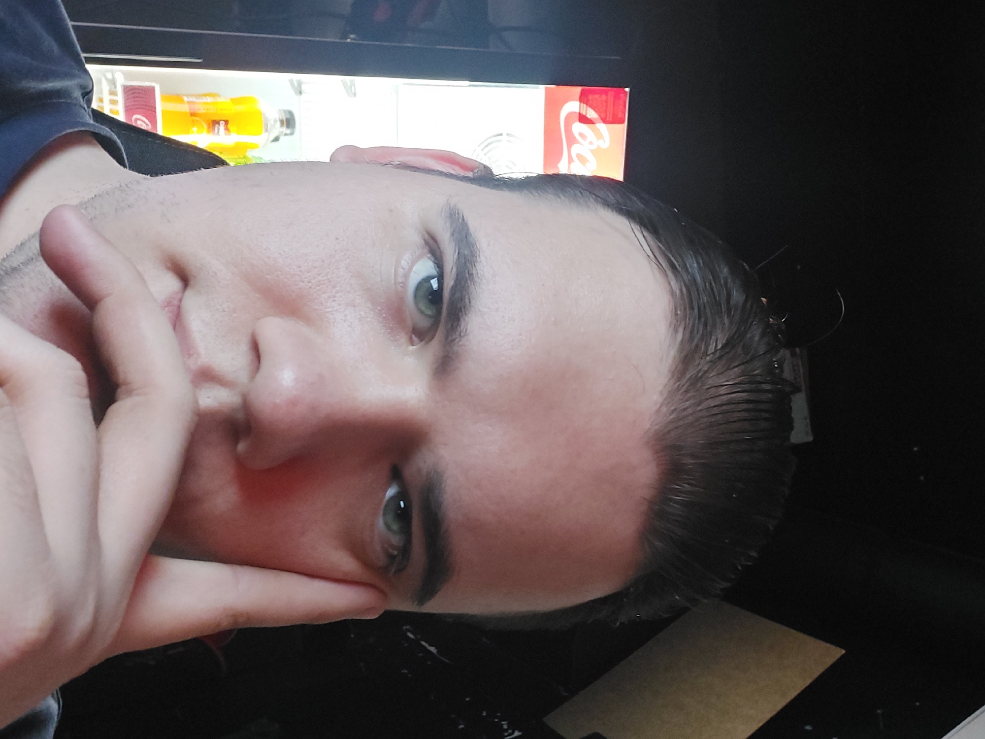 The image captures a close-up, angled portrait of a young male, likely a selfie, taken indoors. The subject's face is oriented diagonally, with his light-colored eyes (appearing green or hazel) looking directly at the camera. His left hand is raised, with fingers resting gently against his chin and cheek, suggesting a thoughtful or casual pose.
The man has dark hair, which appears to be styled back from his forehead, possibly wet or with product. Faint stubble is visible on his jawline and chin. His skin appears clear with a natural complexion.
In the background, behind the man's head, a brightly lit section of what looks like a beverage cooler or refrigerator is visible. An orange-colored drink bottle, with a partially obscured red label that may contain text like "Jamba," is on a shelf. To its right, a red object with the prominent white "Coca-Cola" logo is clearly identifiable, likely a can or a portion of a larger display. The remainder of the background is dark and indistinct, possibly a room at night or with low lighting.
In the bottom right corner, a dark surface, possibly a desk, holds a piece of brown paper or card. Given the context of Tashkent, Uzbekistan, the scene depicts a common indoor setting.