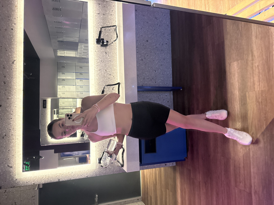 A female individual is depicted standing in a modern fitness facility's changing or lounge area, specifically identified as a "Lounge Zone" within a "SPORTLIFE" gym, located in Kyiv, Ukraine. The image is a mirror selfie, with the subject centered, facing forward, and her legs crossed at the ankles. She has dark hair styled away from her face and is attired in a white sports bra and black athletic shorts, complemented by white athletic shoes. A belly button piercing is visible, and she wears several pink beaded bracelets on her right wrist. Her right hand holds a light-colored smartphone, used for photography, while her left hand holds a clear water bottle with a silver cap, labeled "ARTIC."
The background, visible through the large, rectangular, illuminated mirror, shows a contemporary interior. To the right, a bank of white lockers with black numerical labels (e.g., "1007", "1006", "967") is reflected. A light-colored, speckled countertop runs horizontally below the mirror, upon which two black, wall-mounted hair dryers with coiled cords are situated. A dark door with a silver handle is visible in the reflection to the left. The floor is a dark brown, wood-grain pattern, and a blue rectangular structure, likely a cabinet, stands below the counter on the left side of the frame.