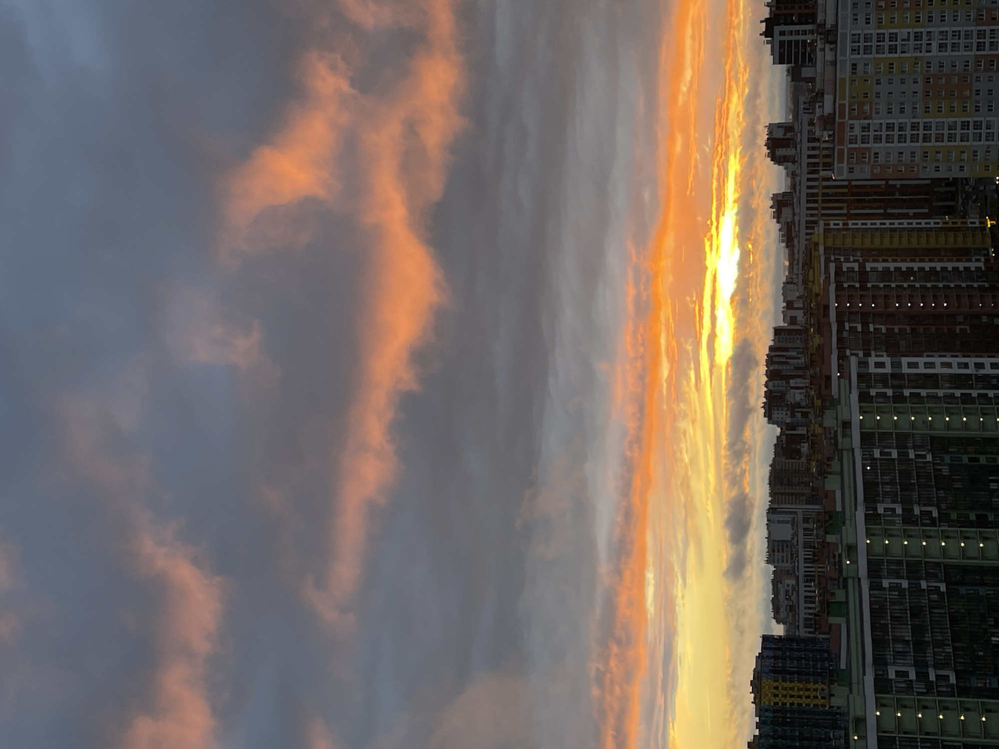 This media file shows a panoramic view of a cityscape at sunset. Tall, modern apartment buildings dominate the right side of the frame, their windows lit, suggesting evening. The buildings are a mix of colors including white, yellow, grey, and dark brown. The left side of the frame is dominated by a dramatic sunset sky. Wispy clouds are illuminated with fiery orange and yellow hues, contrasting with darker grey clouds. The sun itself appears as a bright, blinding light low on the horizon, partially obscured by the clouds. The overall scene evokes a sense of the end of the day in an urban environment. The image is oriented vertically, as if viewed from a high vantage point looking down or across. There is no visible text in the image.