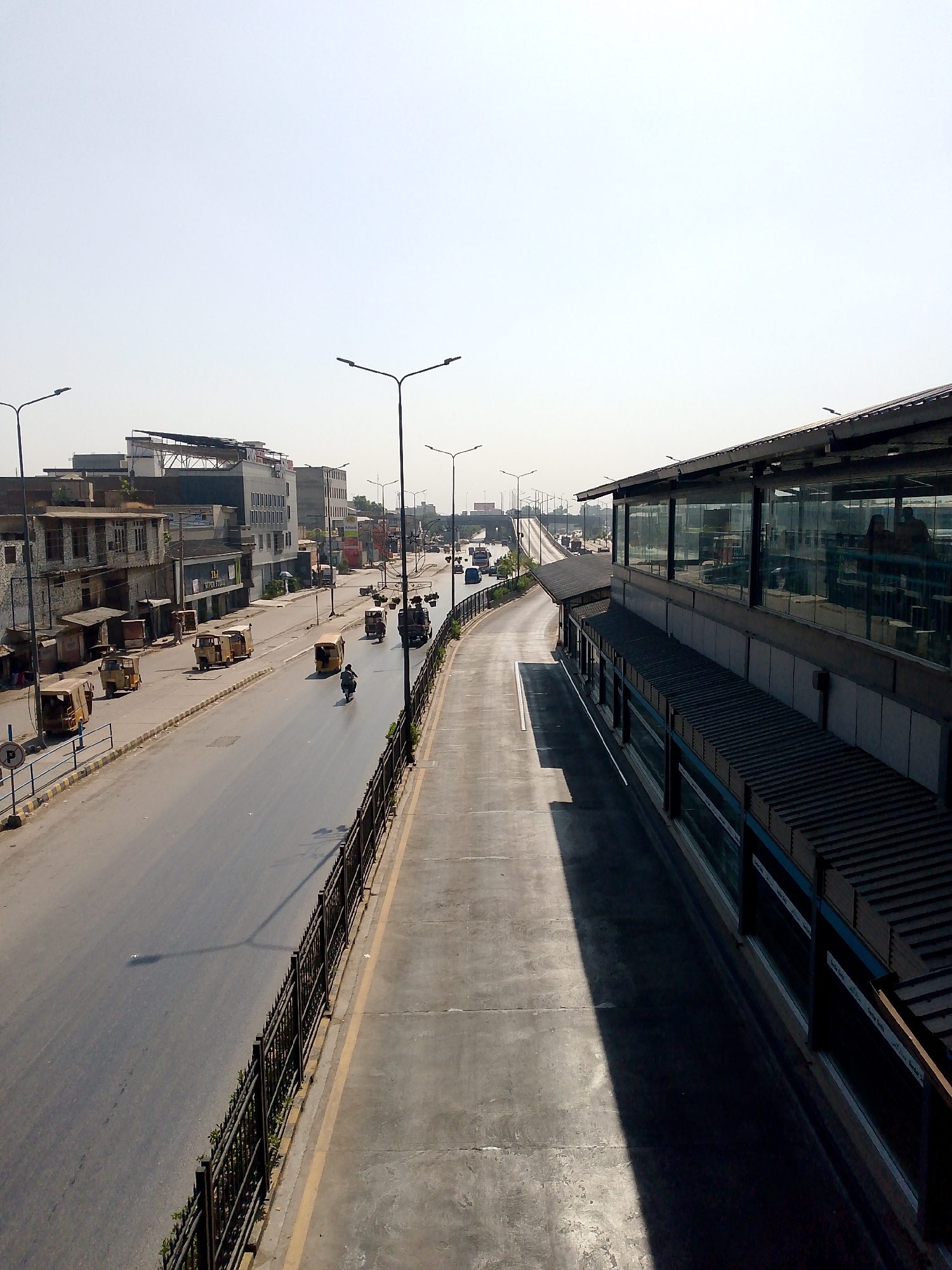 The image depicts an elevated view of a city street and a modern transit station on a clear, sunny day. The street is paved and appears to be a multi-lane road with traffic. Several auto-rickshaws are visible on the left side of the road, some parked and others in motion. A motorcycle rider is seen on the right side of the road. Buildings of varying architectural styles line the left side of the street, ranging from older, weathered structures to more modern commercial buildings. Streetlights are present along the road. On the right side of the image is a contemporary transit station with a long, covered structure featuring a glass-paneled facade and a dark, corrugated roof. The station appears to be elevated above the road. The sky is a bright, pale blue, suggesting daytime. A black metal fence with decorative elements runs along the edge of the elevated walkway or viewing area from which the photograph was taken, separating it from the street below. The scene captures a typical urban environment with ongoing vehicular and pedestrian activity, juxtaposed with modern infrastructure.