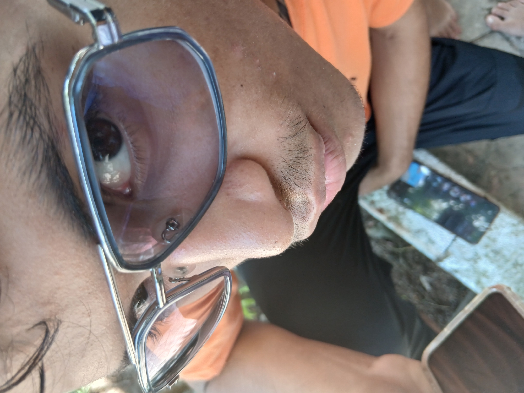 A close-up, tilted perspective shot shows a person with their face turned slightly towards the camera. The person is wearing silver-framed sunglasses that reflect light. The person has dark hair and appears to be wearing an orange shirt. Part of their lap, covered in black pants, is also visible. Next to the person's leg, there's a phone lying on a weathered white surface. Bare feet are seen in the background. Based on the location provided (Playa El Agua, Venezuela), the setting could be outdoors or near the beach.
