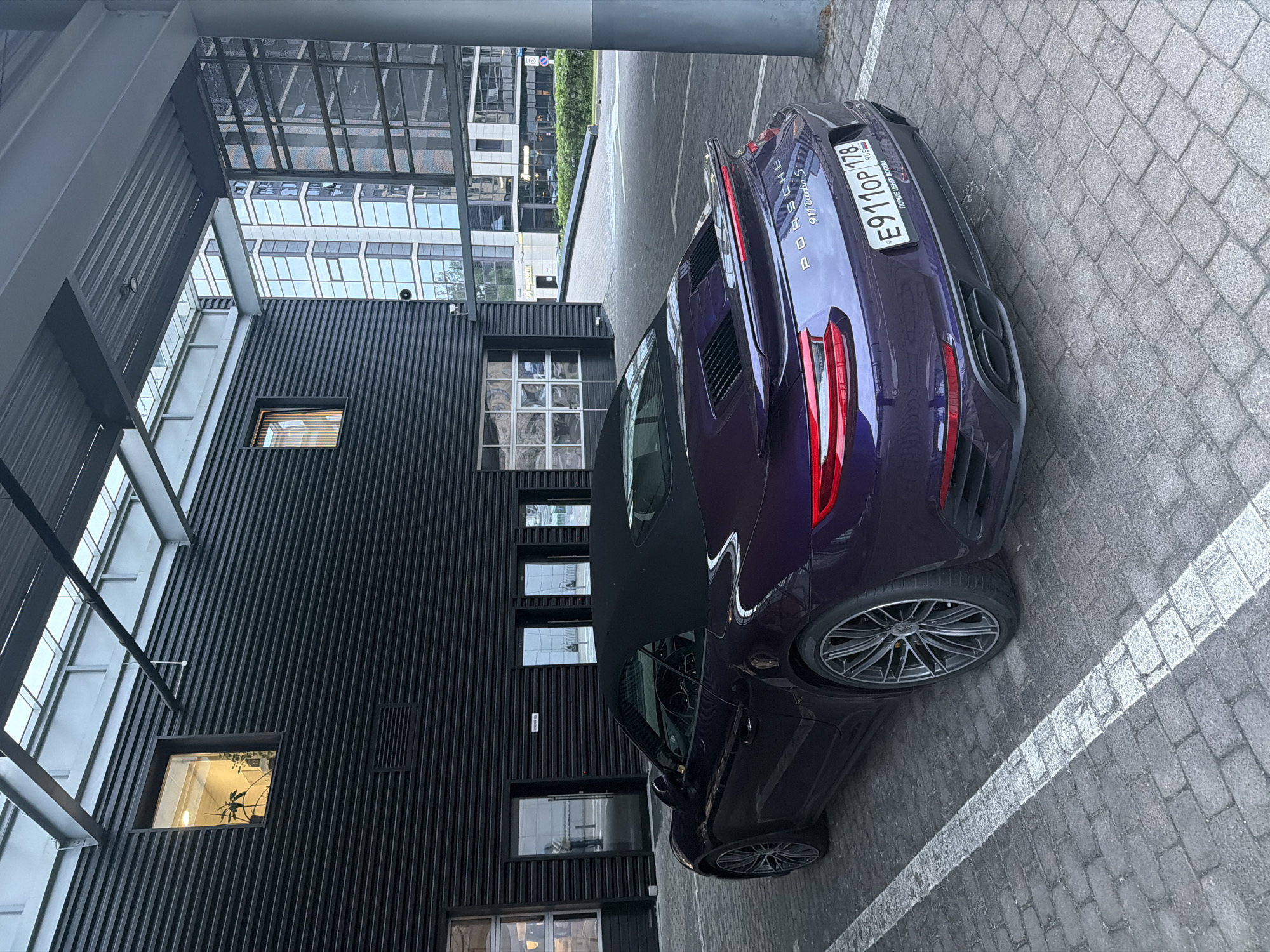 A dark purple Porsche 911 Turbo S convertible is parked in a paved lot. The car is facing away from the camera, with its rear end to the right. The license plate reads "E911OP 178" and "RUS". The car is parked in front of a modern, dark-colored building with horizontal paneling. There are several windows on the building, some illuminated from within. In the background, there are tall, modern glass buildings. The ground is paved with interlocking bricks.