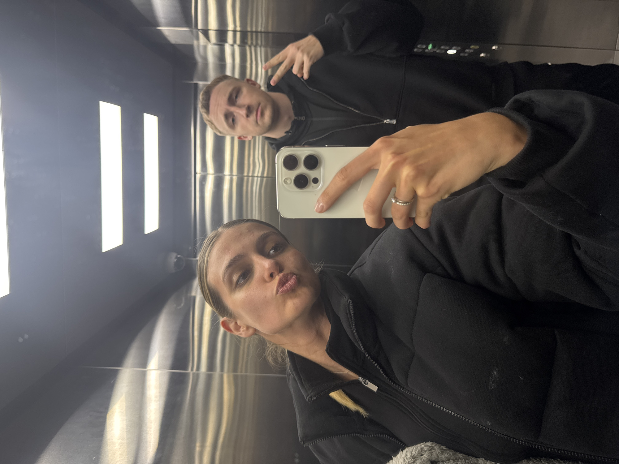 A medium, eye-level shot shows two individuals in what appears to be a mirrored elevator in Unknown, Unknown. A woman, positioned in the lower foreground, is taking a selfie using a white iPhone with three camera lenses. Her face is tilted, and she is puckering her lips for the camera. She is wearing a dark, zip-up jacket over what appears to be a dark top. Her hair is blonde and pulled back. The hand holding the phone has a silver ring on the ring finger.
Behind her and slightly out of focus is a man making a peace sign with his right hand. He is wearing a dark hoodie and has short blonde hair. The elevator has a metallic finish with vertical lines and overhead lighting. The reflection in the mirrored surface shows additional details of the elevator interior.