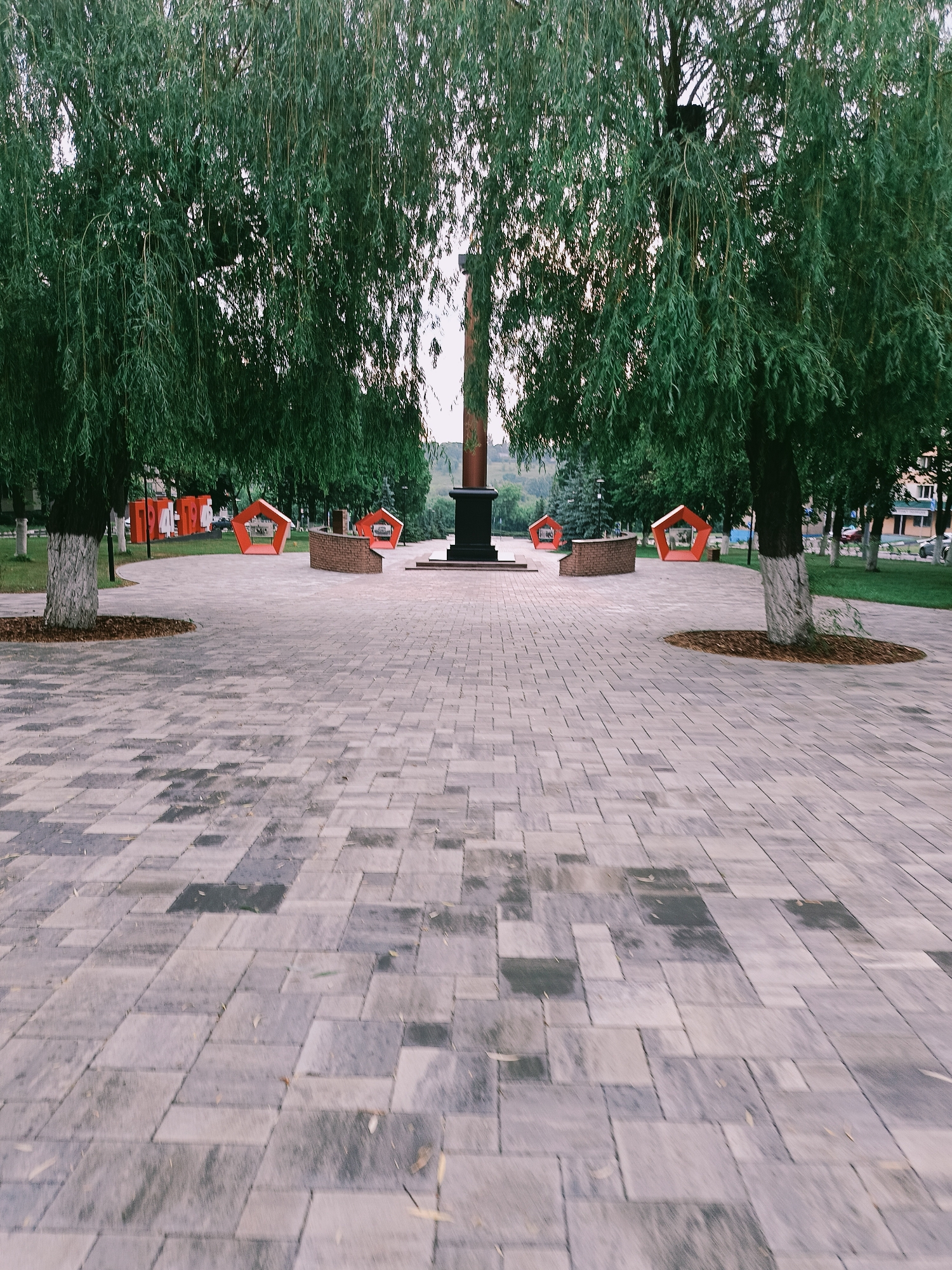 A wide, paved plaza with a mosaic of grey and beige square and rectangular pavers dominates the foreground. The paving extends towards a central monument, a dark, monolithic column atop a tiered base, flanked by two large weeping willow trees with drooping green foliage. To the left and right of the monument, red, pentagonal seating structures with open centers are visible, some set against low brick walls. In the background, beyond the trees and seating, is a grassy area with more trees and buildings. The scene appears to be a public space in the city of Gubkin, Russia, possibly a memorial or park, given the monument and the organized layout. The lighting suggests it is late afternoon or early evening. There are no people visible in the image.