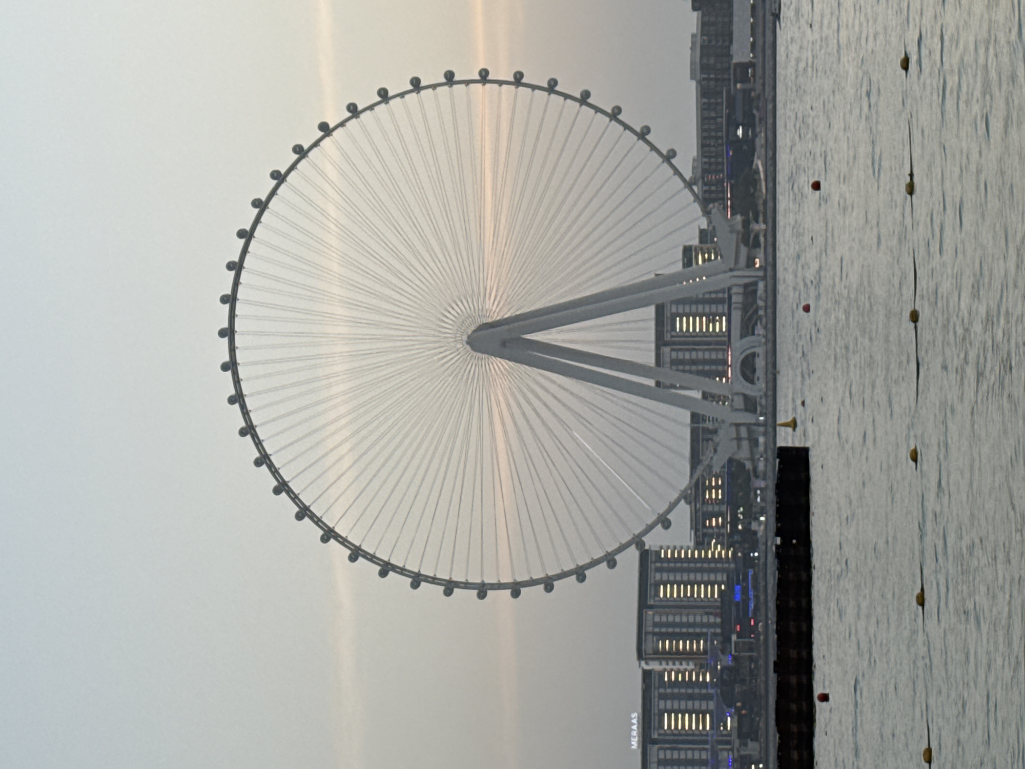 The image captures a wide shot of the Ain Dubai, a giant Ferris wheel, situated in the Jumeirah Beach Residence area of Dubai, United Arab Emirates. The Ain Dubai is centrally located in the frame, its white structure and spokes prominently displayed against a pale, overcast sky. The Ferris wheel is supported by two large, white legs that form an inverted V shape.
In the background, a row of modern buildings lines the waterfront, with lights visible in some windows. The word "MERAAS" is visible on one of the buildings, indicating a development by that company. The foreground consists of a body of water, likely the Arabian Gulf, with a dark, rectangular structure floating on its surface. Several yellow buoys and a few red buoys are scattered across the water, suggesting a designated swimming or boating area. The overall lighting suggests either early morning or late afternoon, with soft, diffused light. There are no people visible in the image.