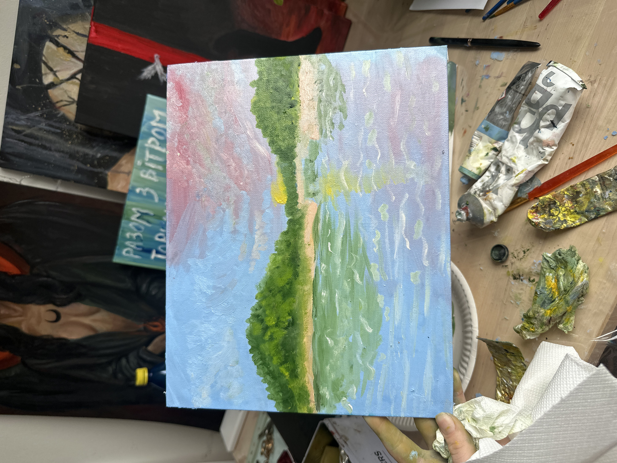 The image depicts a close-up of a painting on an easel, likely in an artist's studio. The painting itself is a landscape scene with a body of water in the foreground and two green, tree-covered hills in the background under a sky with pink and blue hues. The water reflects the sky and hills, with visible brushstrokes indicating it's an oil painting. Surrounding the painting are various art supplies: paint tubes (some crumpled and used), brushes, a palette knife, and crumpled paper towels with paint stains. In the background, other paintings are partially visible, including one with a dark figure and what appears to be a crescent moon symbol. A bottle with a yellow cap is also present. The overall setting suggests an active art creation space. There are no discernible elements that would place this scene specifically in Prague, Czechia.