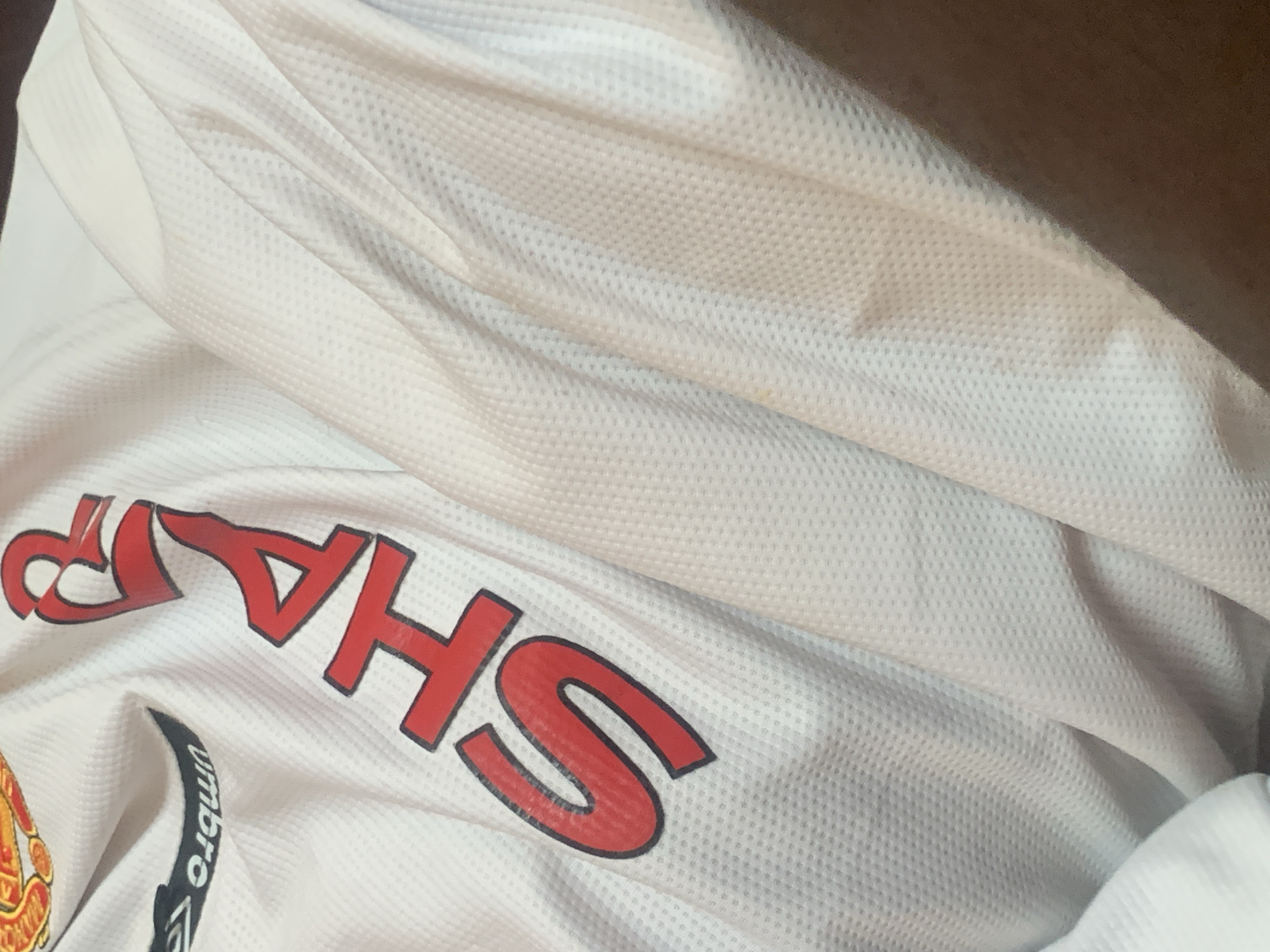 The image shows a close-up of a white Manchester United jersey with red lettering that spells out "SHARP". The jersey has the Umbro logo on the left side of the chest. The fabric of the jersey is textured with small diamond-shaped patterns. The jersey is draped and folded, creating shadows and highlights across its surface. There is a small, faint brownish mark on the fabric to the right of the lettering. The background is a dark brown, out-of-focus area, likely a surface or clothing item. The scene is devoid of any recognizable urban or natural features associated with Jauro Yinu, Nigeria.