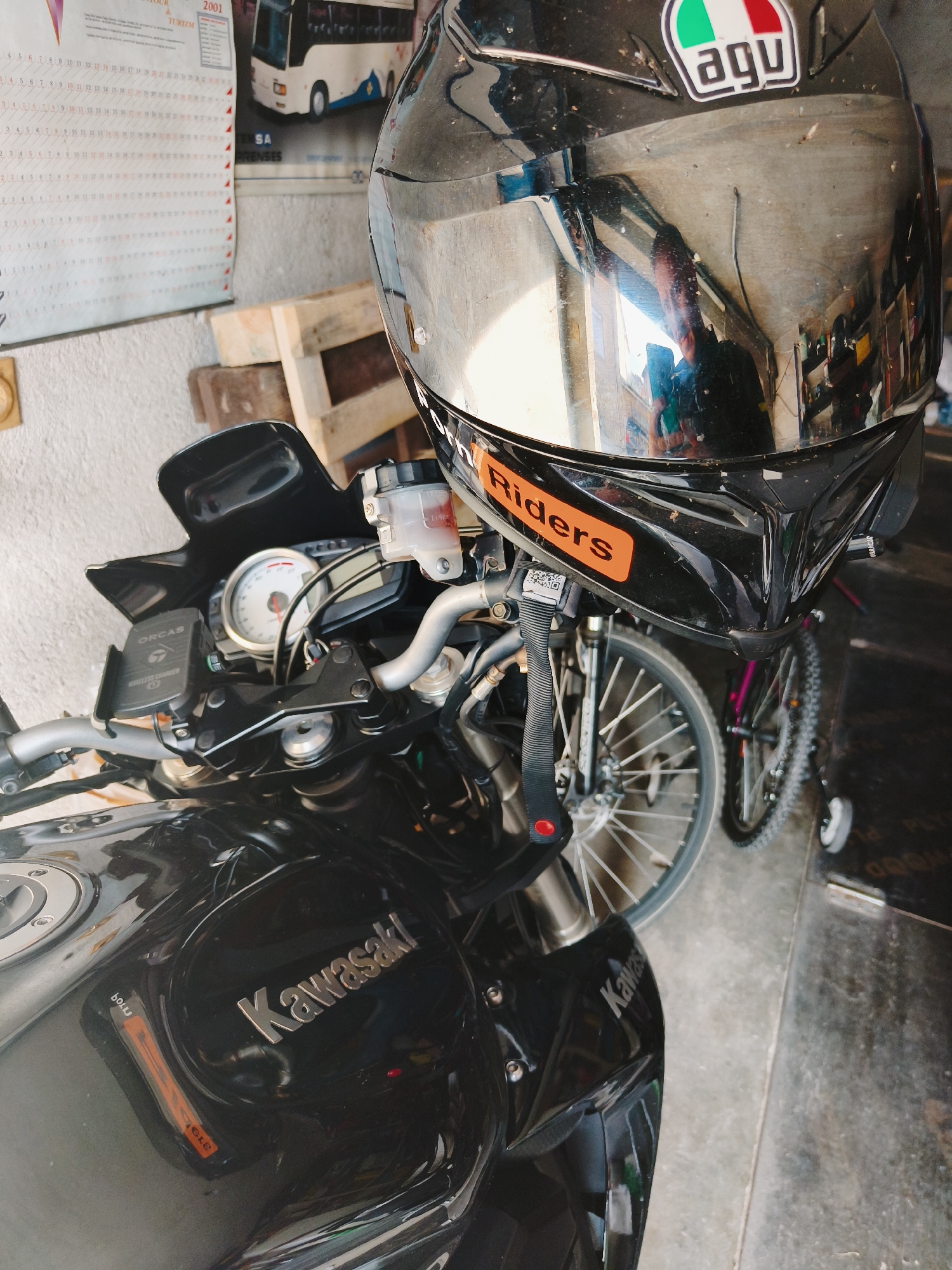 The image shows a black Kawasaki motorcycle with a helmet perched on its handlebars, located indoors. The helmet is black with a reflective visor and an "AGV" logo on top. An orange sticker that reads "Riders" is visible on the front of the helmet. The motorcycle features a dashboard with a speedometer, a black windshield, and a phone holder with the brand "Orcas". "Kawasaki" is emblazoned on the motorcycle's fuel tank. A bicycle is partially visible behind the motorcycle, and a wooden pallet leans against a wall. A calendar with the year "2001" and the words "TOUR & TURIZM" is affixed to the wall. The image was taken inside a room. Bulgaria is mentioned, but its exact location is uncertain from the visible details.