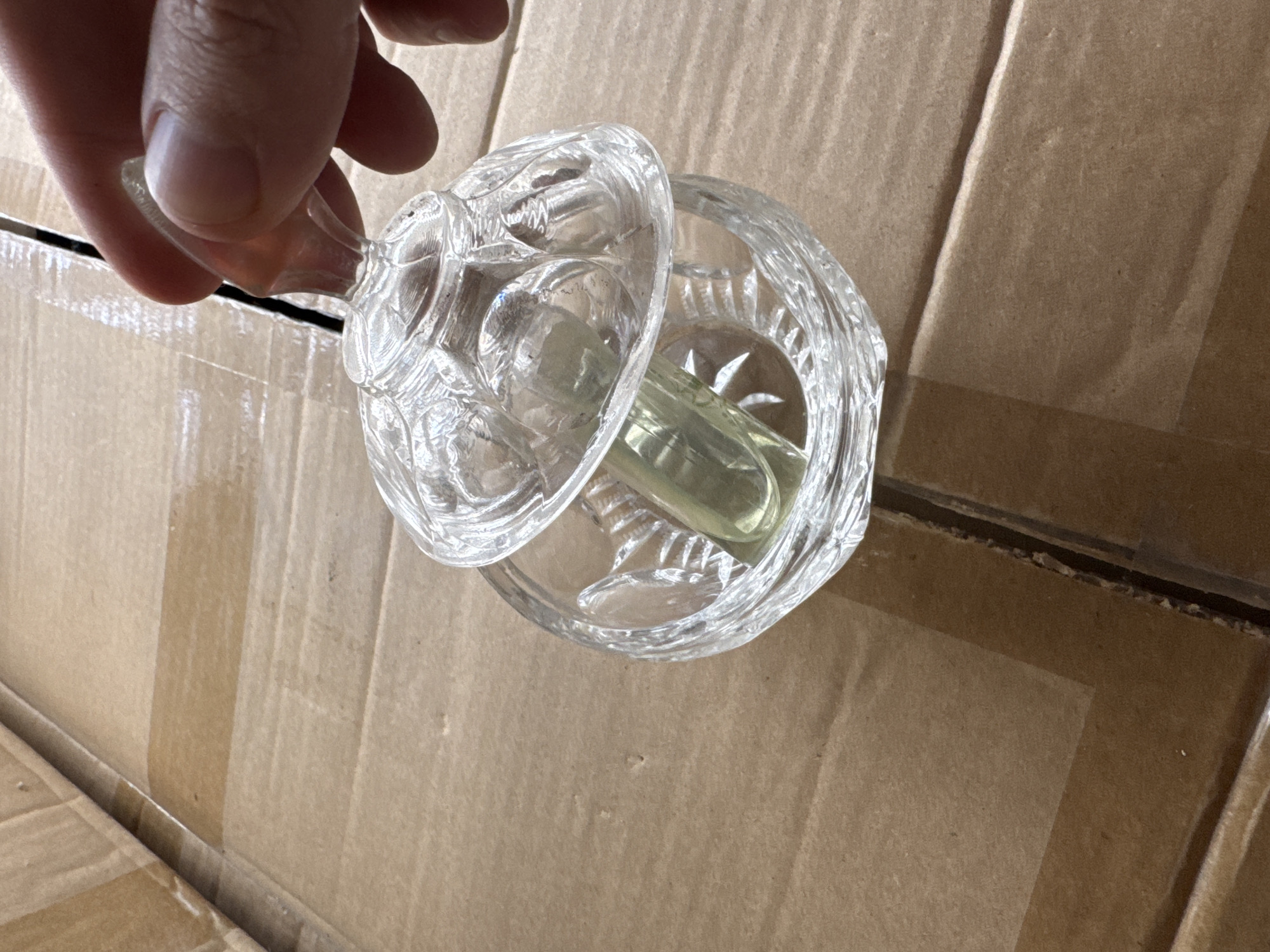 A person's hand is shown holding the lid of a clear, ornate glass jar. The jar contains a light-colored, cylindrical object. The jar is placed on a brown cardboard surface, which appears to be the interior of a shipping box. The scene is lit from above, casting subtle shadows on the cardboard. The image does not provide any specific information about the location in the city of Rozdilna, Ukraine.