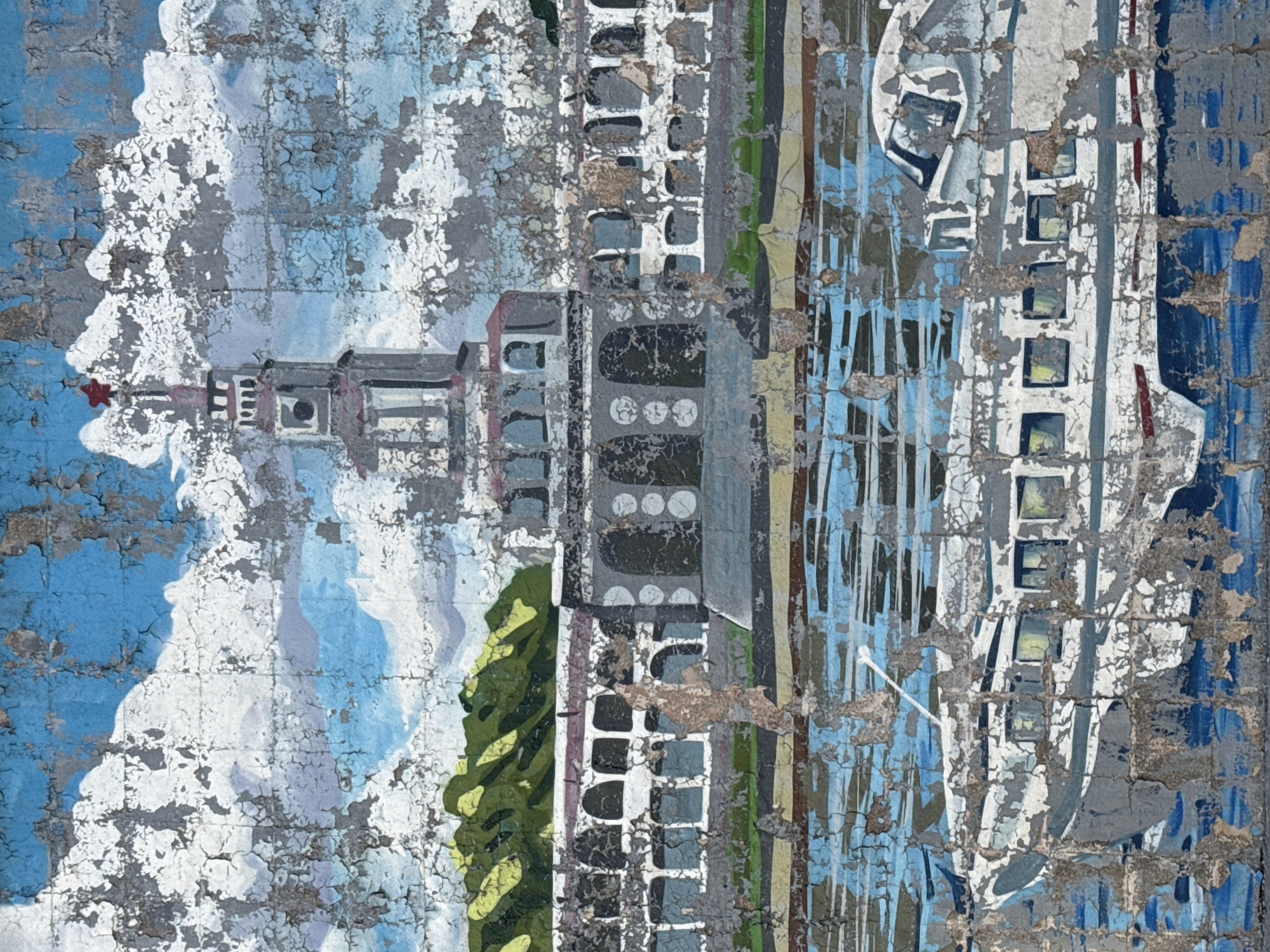 The image depicts a weathered mural or painting that appears to be a stylized scene of Moscow, Russia. On the left, there is a building with a distinctive tower topped with a red star, reminiscent of one of the Seven Sisters skyscrapers built during the Stalinist era, specifically Lomonosov Moscow State University. Adjacent to this, the mural presents a row of buildings with repeated window patterns and architectural elements. Green foliage is visible near the buildings, suggesting trees or parkland along a riverbank. To the right, the image includes a large, white ship, potentially a river cruise vessel, which is rendered in a simplified style. The background features a blue sky with white clouds, and the overall scene seems to be located along the Moskva River. The mural itself exhibits significant signs of wear and tear, with the paint peeling and flaking off, revealing the surface beneath.