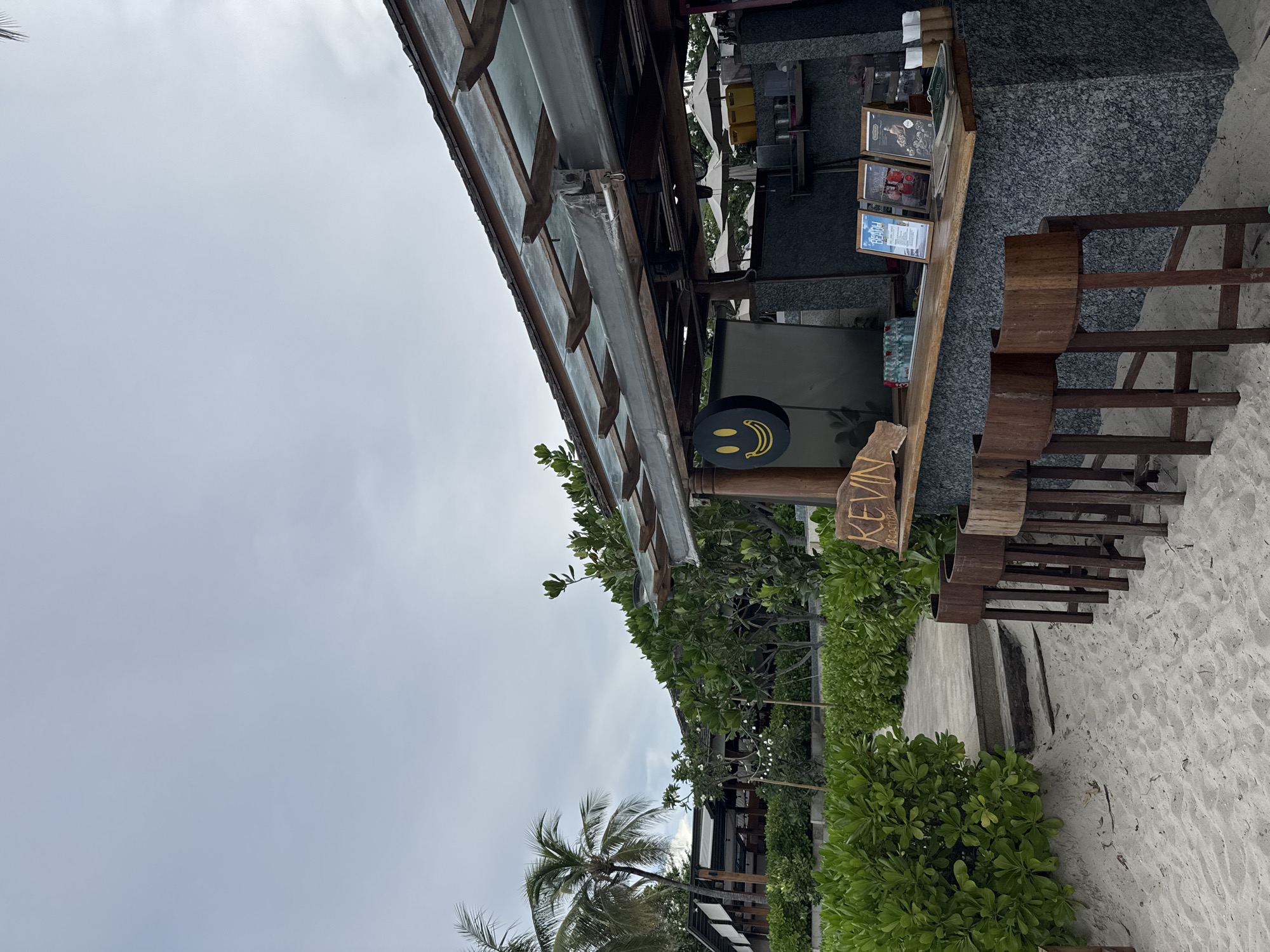 An outdoor beach bar named "KEVIN Beach Bar" is depicted under a cloudy sky. The bar features a granite counter with three framed menus and several bottles of water and soda. Four wooden bar stools with curved seats are placed in front of the counter on the sandy beach. A circular sign with a yellow smiley face is hanging on the left side of the bar. Lush green bushes line the pathway leading to the bar, and palm trees are visible in the background. A building with a modern architectural style, characterized by a wooden structure and a glass roof, is situated behind the bar area. The scene suggests a relaxed atmosphere at a beachside establishment.