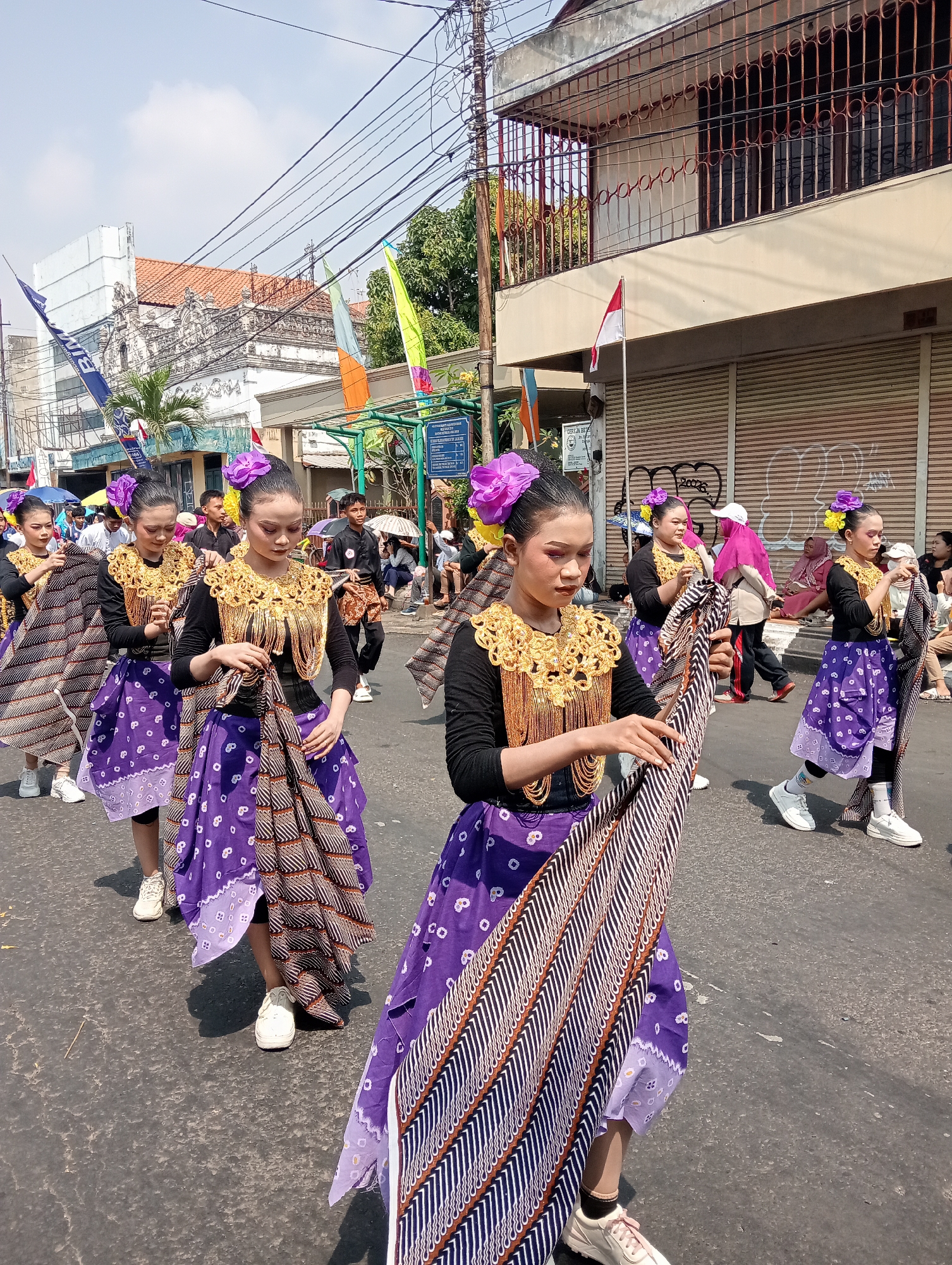 In Bintoro, Indonesia, a group of young women participate in a street parade, dressed in traditional Indonesian attire. The women, with their hair styled in buns and adorned with large purple flowers, wear black long-sleeved shirts and vibrant purple skirts. Their outfits are accessorized with ornate gold necklaces. Each woman carries a patterned cloth, which they are either holding or adjusting. They are walking in a line on a paved street under a clear, sunny sky. In the background, buildings with red-tiled roofs and white facades line the street. Some buildings have metal grates on their windows, and one has a flag flying. Spectators are visible on the sidewalks, some holding umbrellas. Power lines crisscross the sky above the scene.