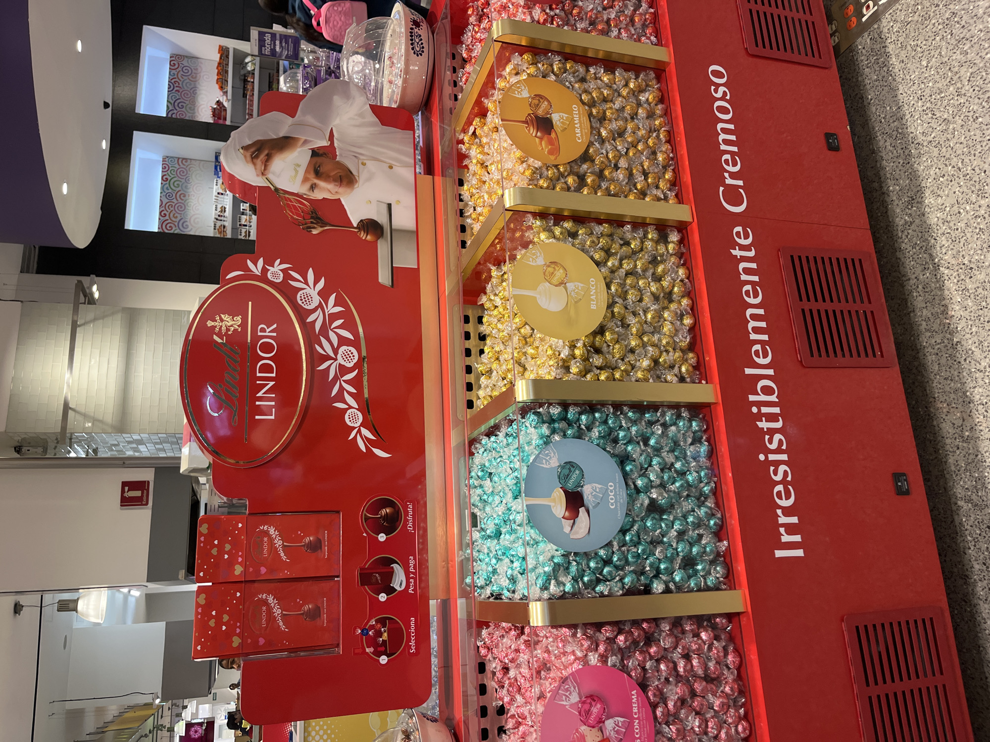 The image shows a Lindt Lindor chocolate display stand in a store. The stand is red and features images of a chef and the Lindor logo. The chocolates are arranged in four tiers, each containing a different flavor: caramel, white chocolate, coconut, and cream. The side of the stand has the text "Irresistiblemente Cremoso" written in Spanish. Based on the presence of Spanish text and signage, it is likely this display stand is located in a store in Puebla, Mexico.