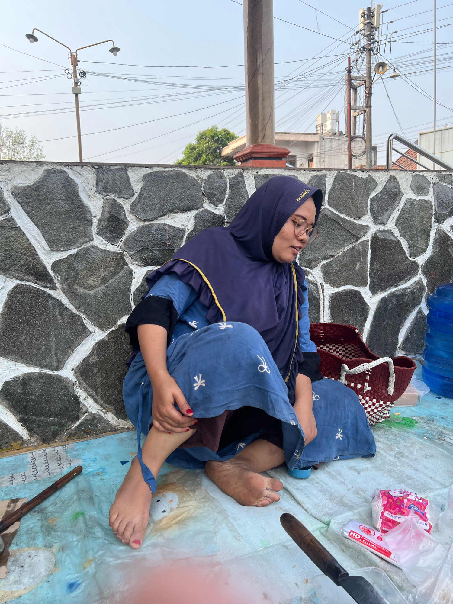 A woman wearing a navy blue hijab and a matching blue patterned dress is sitting cross-legged on a light blue mat outdoors. She has glasses and appears to be speaking. To her right, a woven basket and a large blue water cooler are visible. In the foreground, a spatula or knife with a wooden handle lies on the mat, along with a package of tissues and a plastic container. The background features a stone wall with white mortar, a street lamp, power lines, and buildings. The lighting suggests it is daytime, possibly late afternoon. The location is identified as Kecamatan Pati, Indonesia.