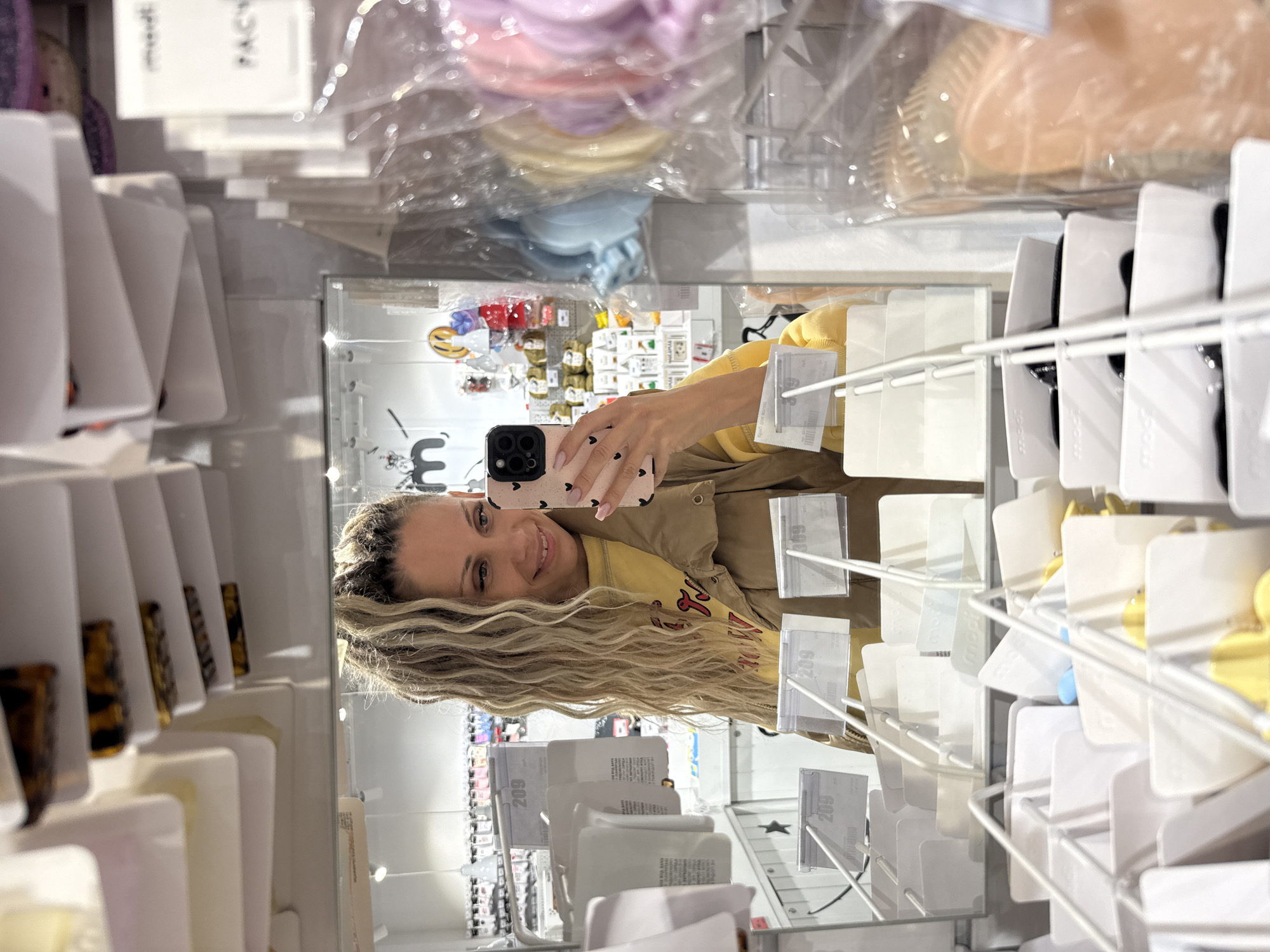 The image shows a woman taking a mirror selfie in a store. She has long blonde hair styled in dreadlocks and is wearing a yellow sweatshirt and a tan jacket. She is holding a pink phone case with black hearts. The background reveals shelves filled with various hair accessories, such as hair clips and scrunchies, as well as other small retail items. The store appears to be a beauty or accessories shop. The image does not contain any information indicating it is located in Yaroslavl, Russia.