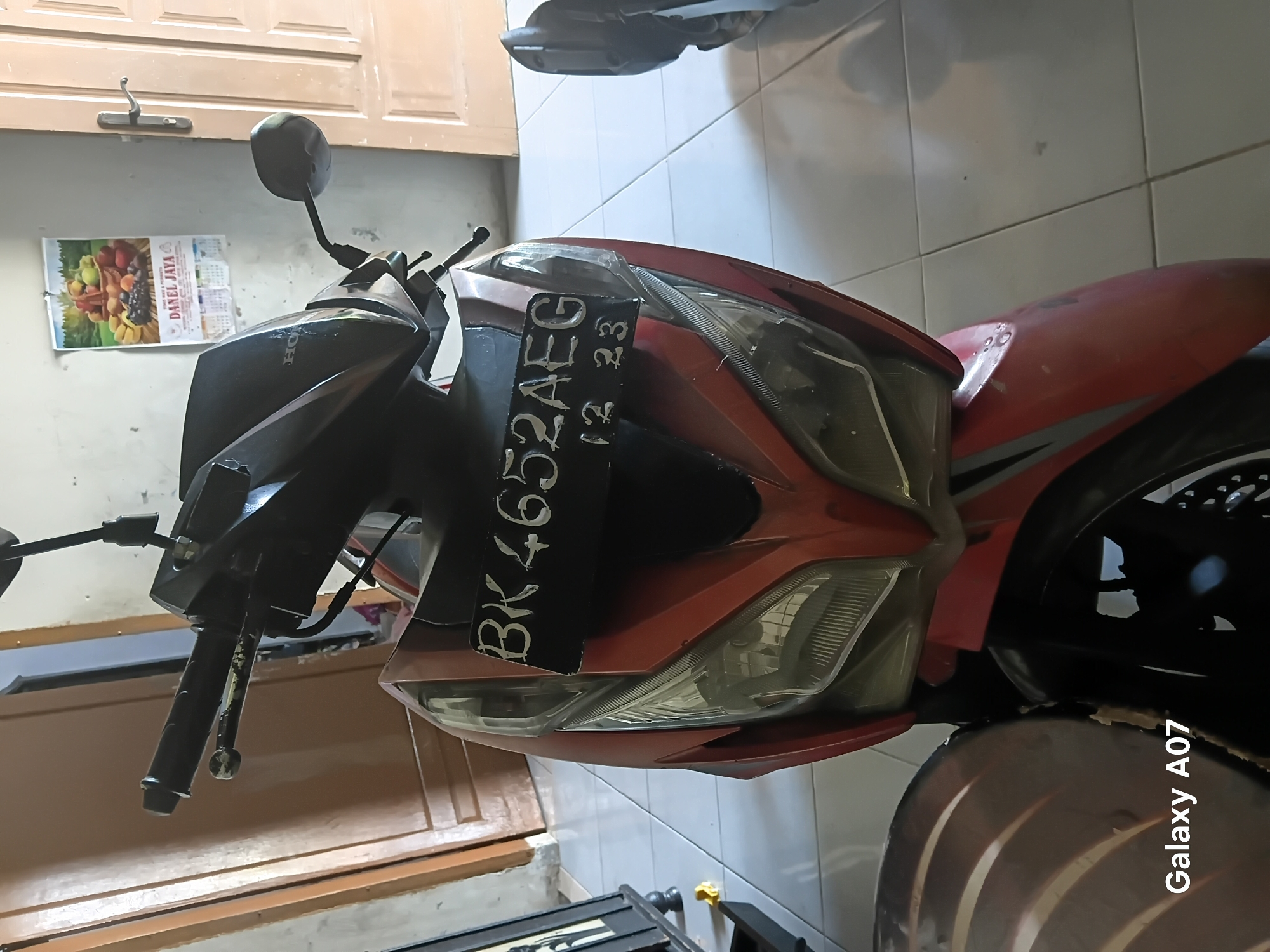The image shows the front of a red and black motorcycle parked indoors, likely in a garage or a residential area in Tembung, Indonesia. The motorcycle has a license plate that reads "BK 4652 AEG 12 23". The background includes a beige door on the right and a wooden door on the left. A calendar with an image of fruits is hanging on the wall behind the motorcycle. The floor is tiled in a light color. The lower left corner of the image has a watermark that says "Galaxy A07".