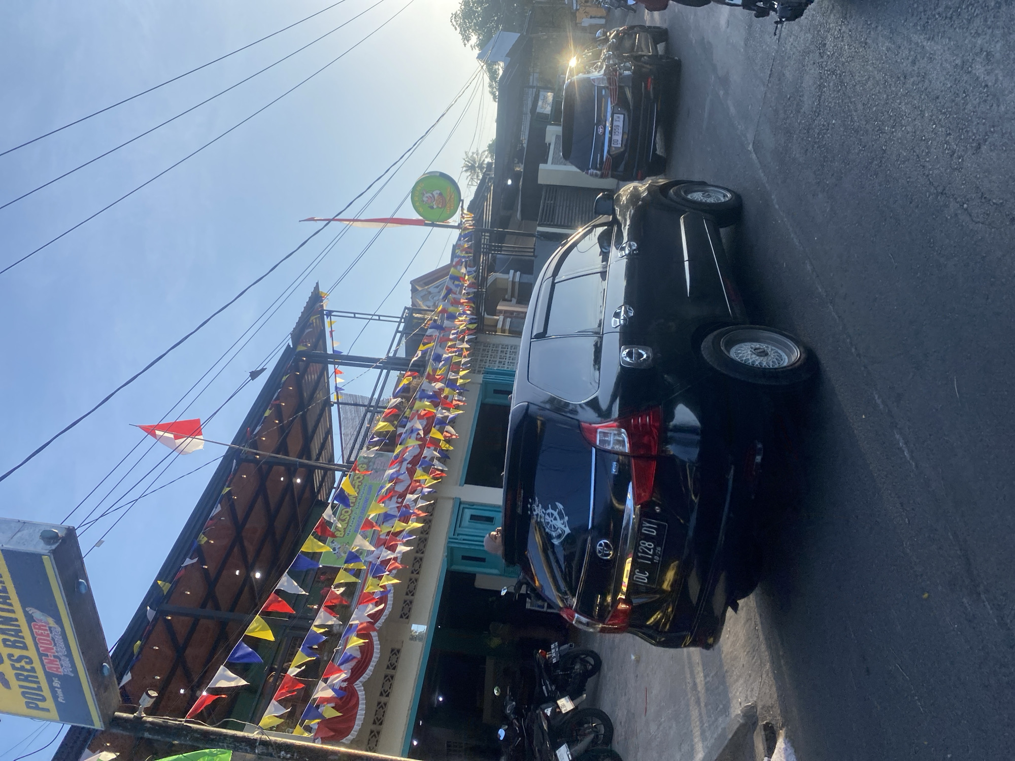 The image shows a street scene in Bantaeng, Indonesia, likely near a police station given the visible sign for "POLRES BANTEN". The foreground is dominated by the rear of a black MPV with license plate "DC 1128 DY". To the left, several motorcycles are parked. Behind the vehicles, a building with an awning decorated with colorful triangular flags and red and white ruffled fabric is visible. A sign for "BAKSO PAK ARIS" is also present, indicating a food establishment. An Indonesian flag flies from a pole near the building. The sky is clear and blue, with the sun casting long shadows, suggesting it is late afternoon or early morning. Another black MPV is visible further down the street, and a pedestrian can be seen entering the building. The environment appears to be a commercial or mixed-use area on a street.
