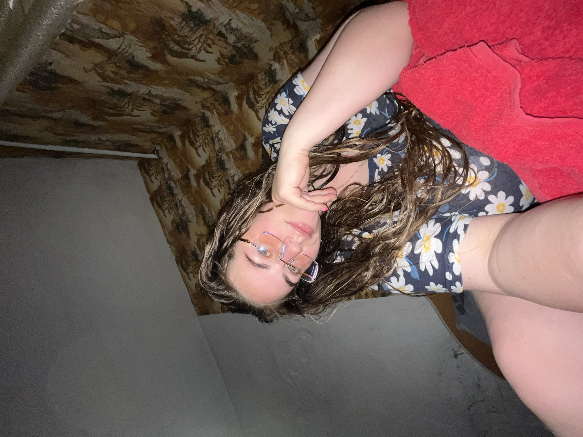 An interior scene in Сосново, Russia, features a young woman with long, wavy brown hair, wearing a dark blue short-sleeved top adorned with white and yellow daisy patterns. She is positioned facing the camera, resting her left hand under her chin, and is wearing rimless or thin-rimmed glasses that reflect light, creating a subtle pink/orange hue on the lenses. Her fingernails have pink polish. A bright red, textured fabric, likely a blanket or towel, is partially draped over her lower body on the right side of the frame. The background consists of a plain, light grey textured wall on the left and a wall covered with patterned wallpaper on the right, depicting a savannah landscape with acacia-like trees and zebras in shades of brown, beige, and dark green. A thin white vertical pipe or conduit runs against the patterned wallpaper. Above, a plain grey textured ceiling is visible, with a corrugated silver pipe or vent running along the upper right corner where the wall and ceiling meet.