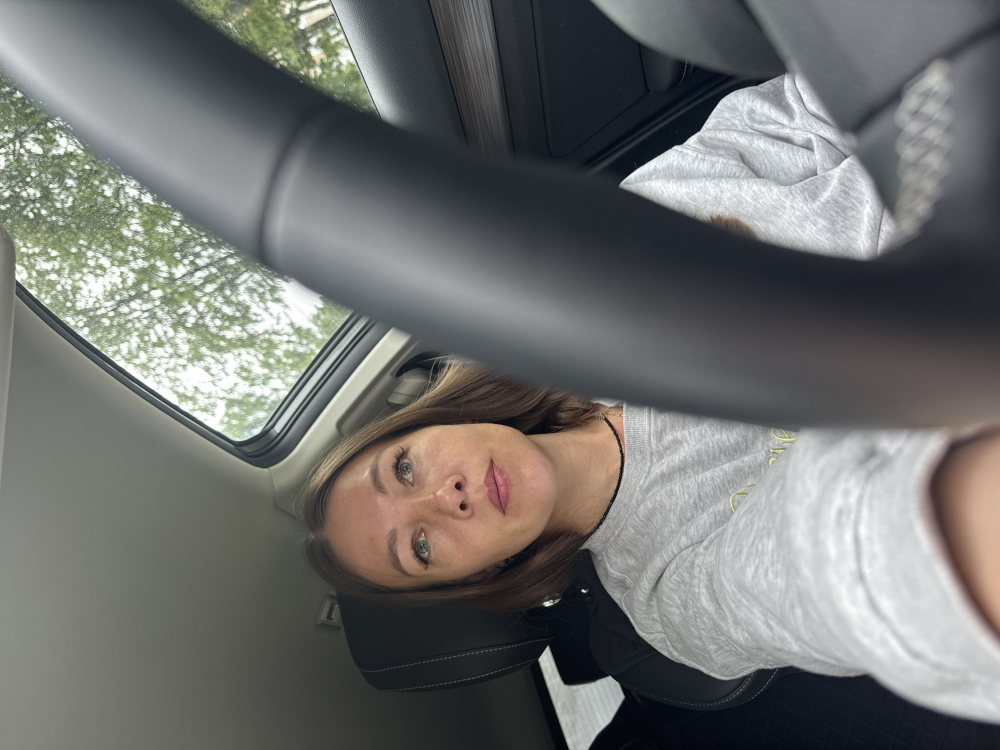 A medium shot shows a woman sitting in the driver's seat of a car. She is looking up and to the right, with her eyes slightly open and her mouth closed. The woman has brown hair and is wearing a gray sweatshirt and a black beaded necklace. The car's steering wheel is visible in the foreground, slightly out of focus. The sunroof is open, allowing natural light to illuminate the interior of the car. Trees and foliage are visible through the sunroof and the car's windows, suggesting an outdoor setting. The background is blurred, but the interior of the car, including the dashboard and seats, is discernible. The image appears to have been taken on a cloudy day.