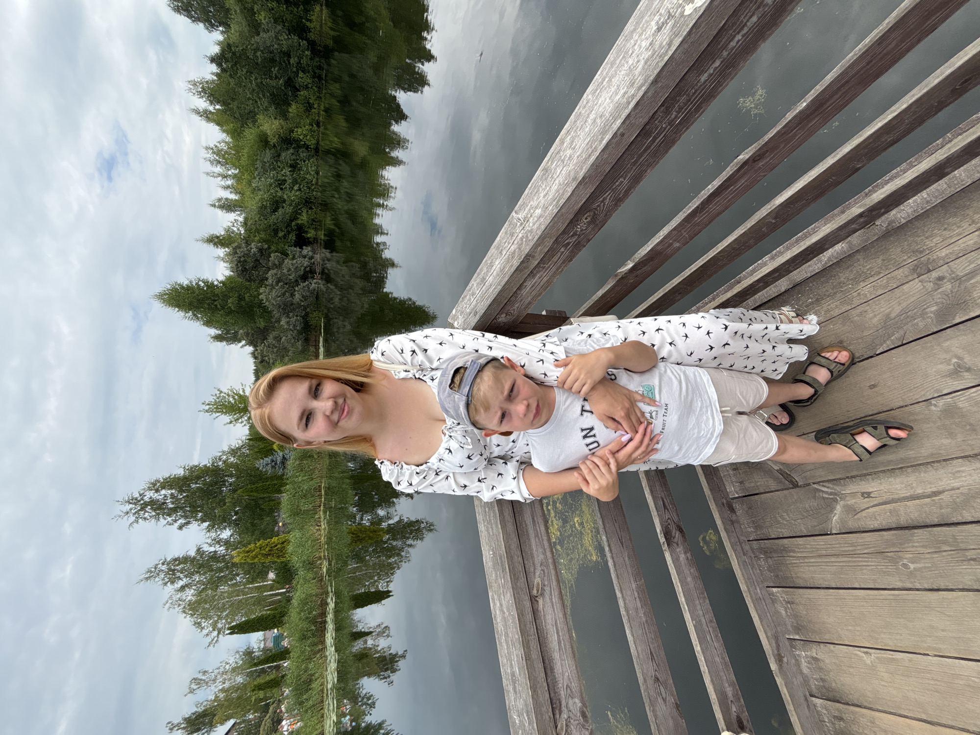 A woman with blonde hair and a white dress with a bird pattern smiles at the camera, her arm around a young boy wearing a white t-shirt and a grey baseball cap. The boy is looking downwards, with his hand resting on his stomach. They are standing on a wooden pier next to a body of water. The water reflects trees and the sky. The sky is cloudy, suggesting it might be an overcast day. The scene appears to be outdoors, possibly in a park or by a lake. The presence of trees and water suggests a natural setting. The overall mood seems relaxed and happy, with the woman's smile and the casual poses of both individuals. The image is oriented vertically. There is no visible text in the image.