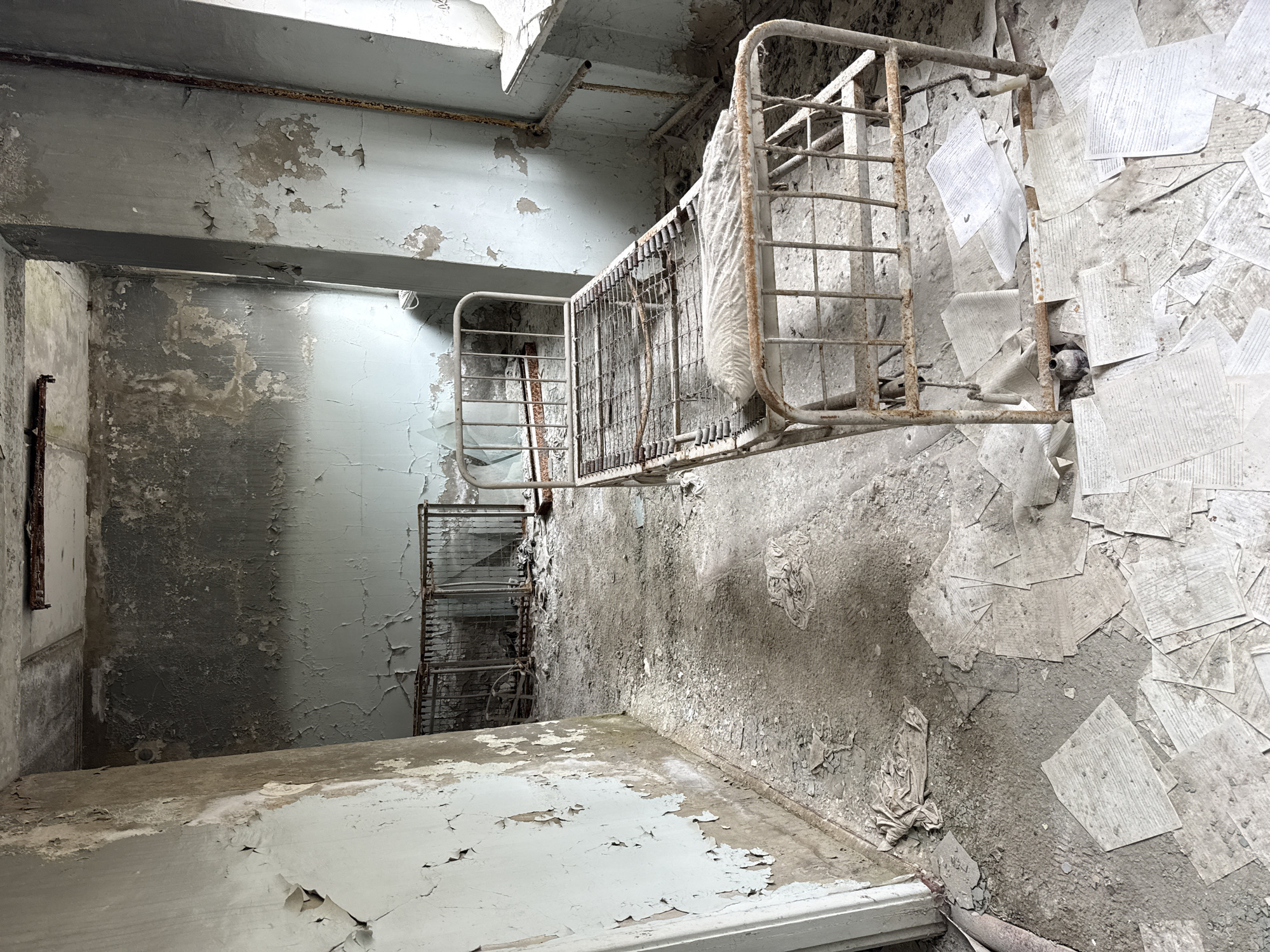 The image depicts an interior room in an abandoned building, possibly in Pryp'yat', Ukraine. The walls and ceiling exhibit significant peeling paint and deterioration. A metal-framed bed, also showing signs of rust and decay, is partially visible, with what appears to be a stained, dusty mattress. The floor is covered in debris, including scattered papers. Natural light enters from an unseen source. The room is in a state of severe disrepair and dereliction.