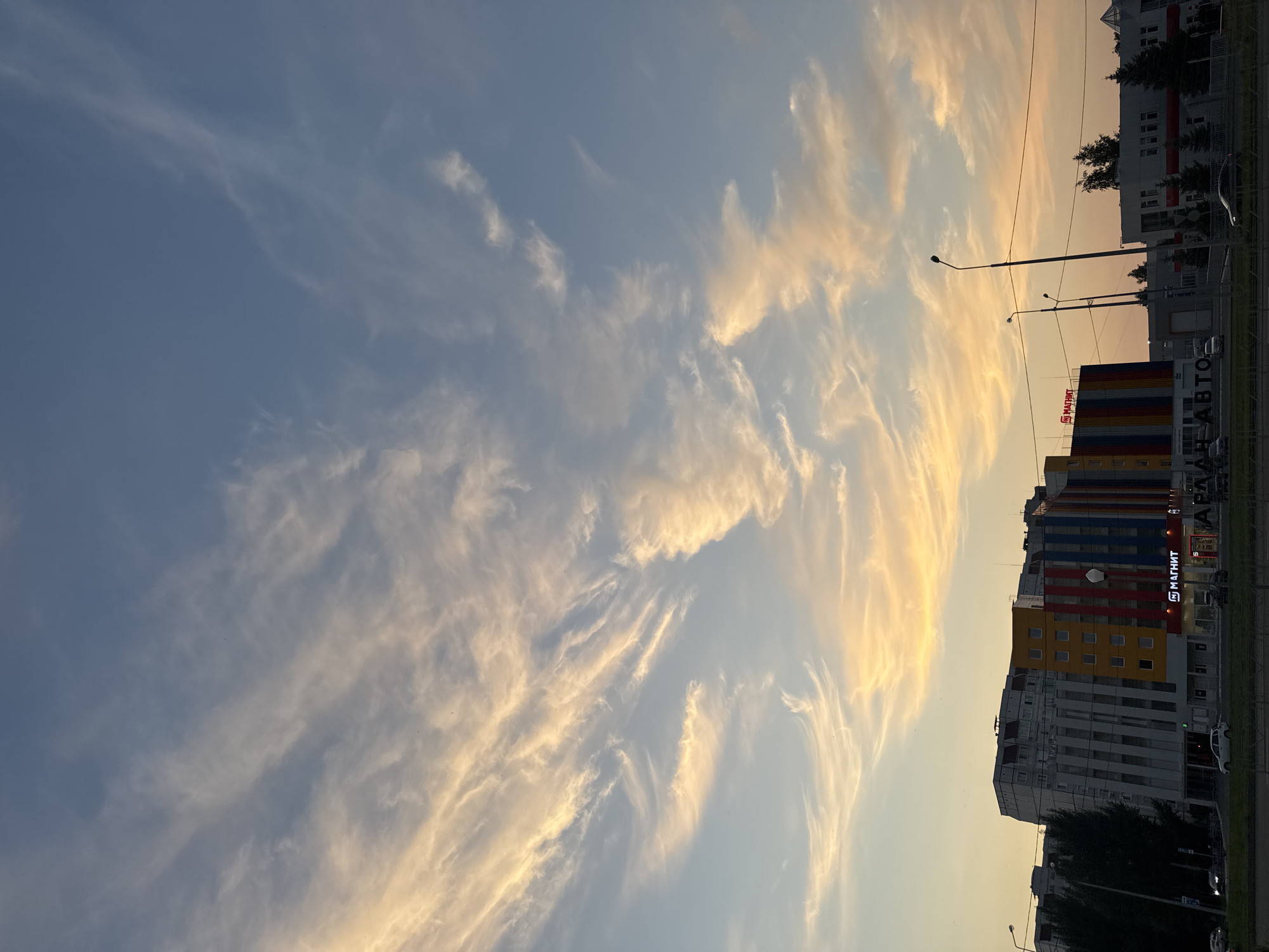 The image captures a street view in Tolyatti, Russia, at dusk. The sky is filled with wispy, golden clouds illuminated by the setting sun, contrasting with the deepening blue above. In the midground, a modern building with a colorful facade, featuring sections of yellow, red, and blue stripes, stands prominently. A sign for "МАГНИТ" (Magnit), a Russian supermarket chain, is visible on this building. To the left, another building with a grey and white facade is partially obscured by trees. A street with cars driving on it is in the foreground, with streetlights and power lines stretching across the scene. The overall atmosphere is one of a typical urban evening.