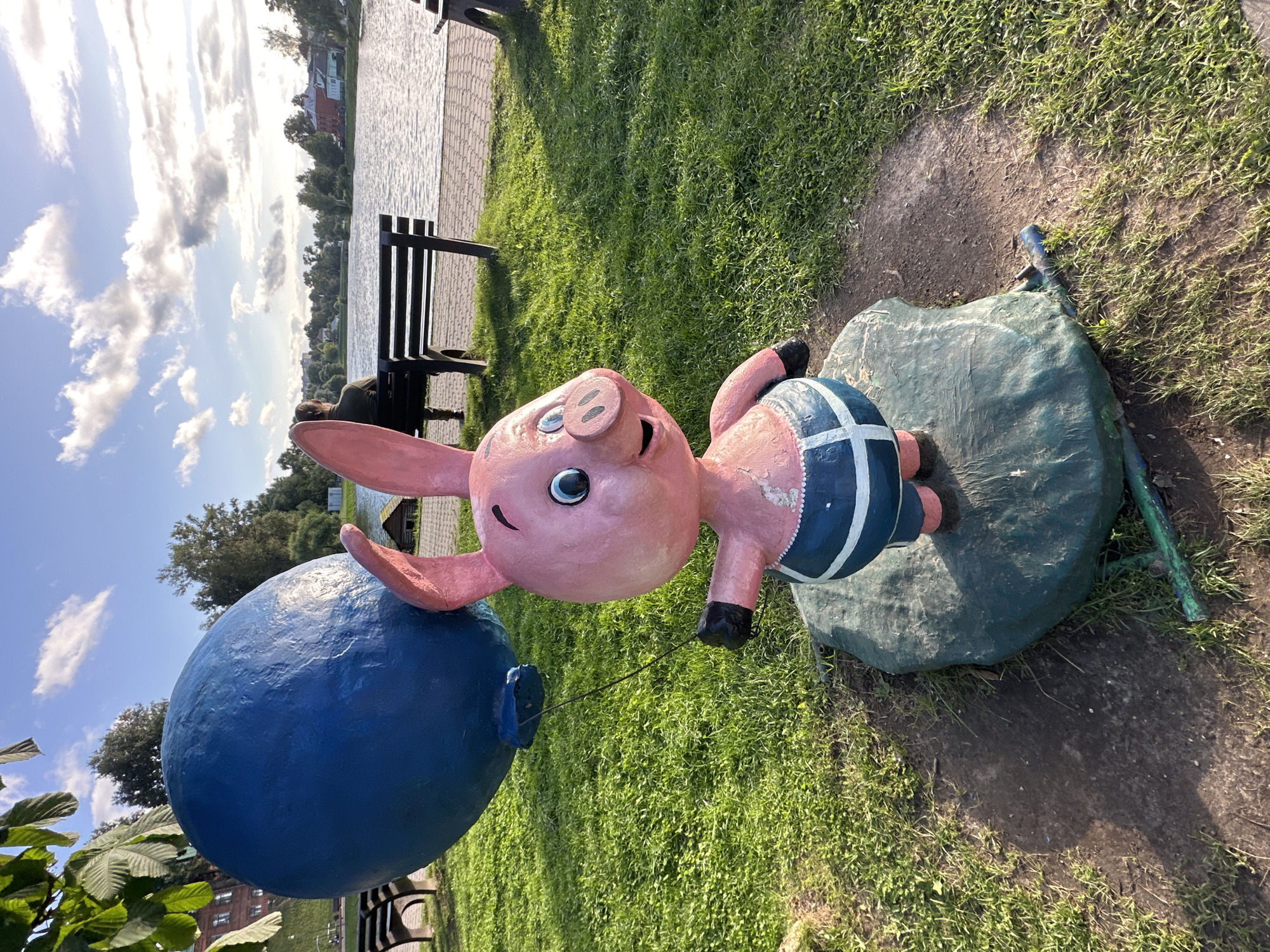 A whimsical pink pig sculpture holding a large blue balloon stands in a park in Sergiyev Posad, Russia. The pig, with big blue eyes and a smiling snout, wears blue shorts with white stripes. It is mounted on a green, rock-like base. In the background, a serene body of water, likely a river or lake, reflects the sky. A paved walkway lines the water's edge, with park benches positioned along it. One bench is occupied by a person sitting with their back to the camera, observing the water. Trees line the opposite bank, with buildings visible in the distance. The scene is bathed in the soft light of a late afternoon or early evening sun, casting shadows on the well-maintained green lawn.