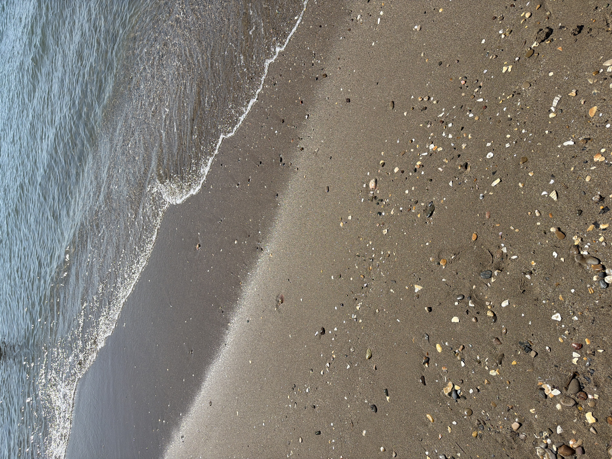 This is an overhead, high-angle shot of a sandy beach with gentle waves lapping at the shore. The water, on the left side of the frame, is a muted blue-gray and shows rippling patterns from the movement of the waves. The edge of the water transitions to wet sand, which then gives way to drier, darker sand speckled with small pebbles and shell fragments. The tide appears to be going out or is very low, as a clear line of wet sand marks the water's edge. The light suggests it is daytime, possibly late afternoon due to the long shadow cast by the wave foam on the sand, indicating the sun is at a relatively low angle. The overall impression is one of a serene, natural coastal environment, likely located in Sérignan, France, given the context. There is no visible text in the image.