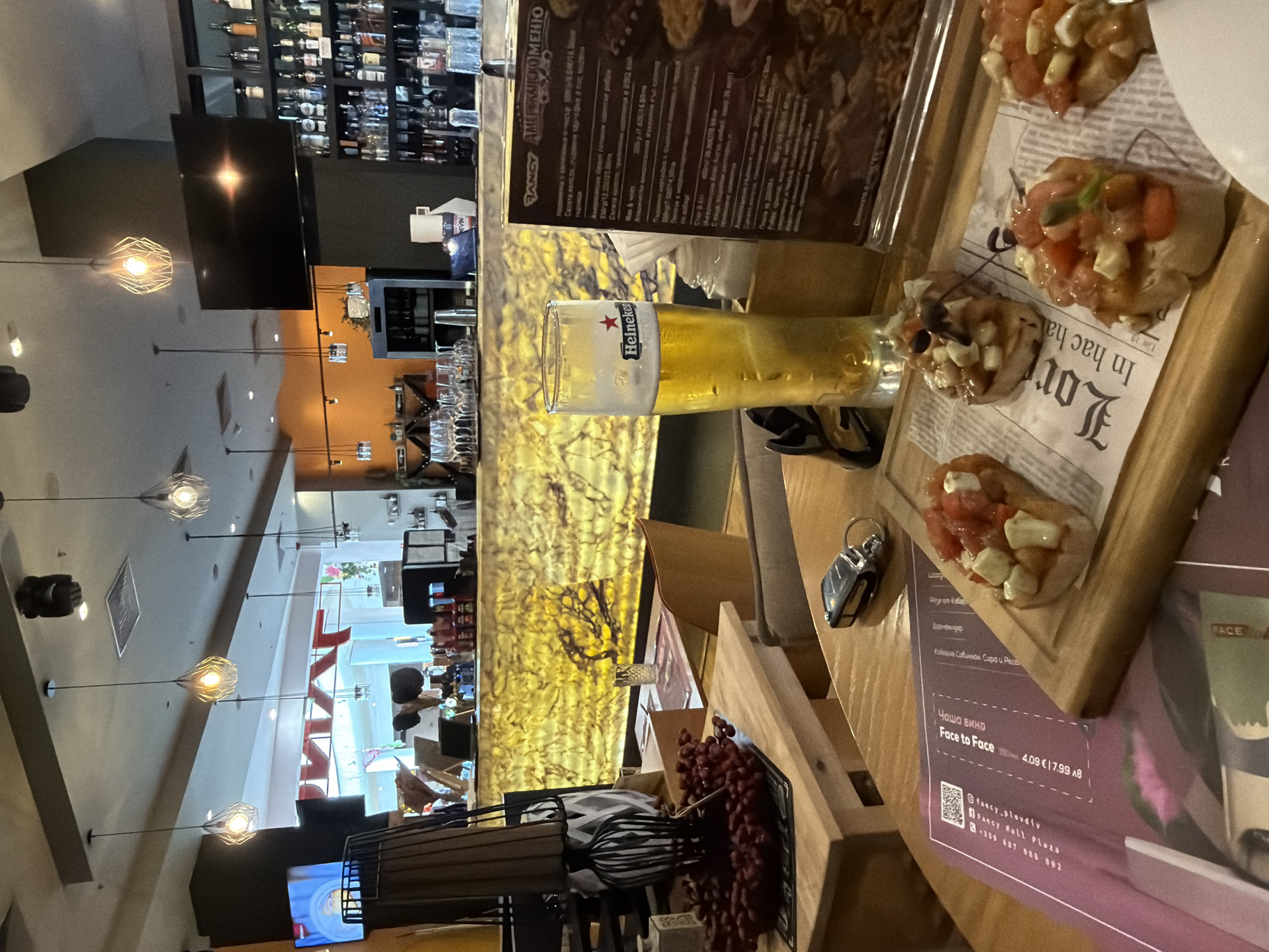 The image depicts the interior of a bar/restaurant located in Plovdiv, Bulgaria (as indicated by the menu mentioning "Fancy Plovdiv"). A Heineken beer and a wooden tray of bruschetta are on the table. The interior includes a bar stocked with bottles. The location seems to be in a mall, as indicated in the menu ("Fancy Mall Plaza"). The lighting fixtures are decorative pendant lights. The background shows people walking inside the mall. There is also a menu that partially covers a menu with drink prices, including a "Face to Face" wine for €4.09. An umbrella and some decorations are on the table next to the food. A key is near the bruschetta.