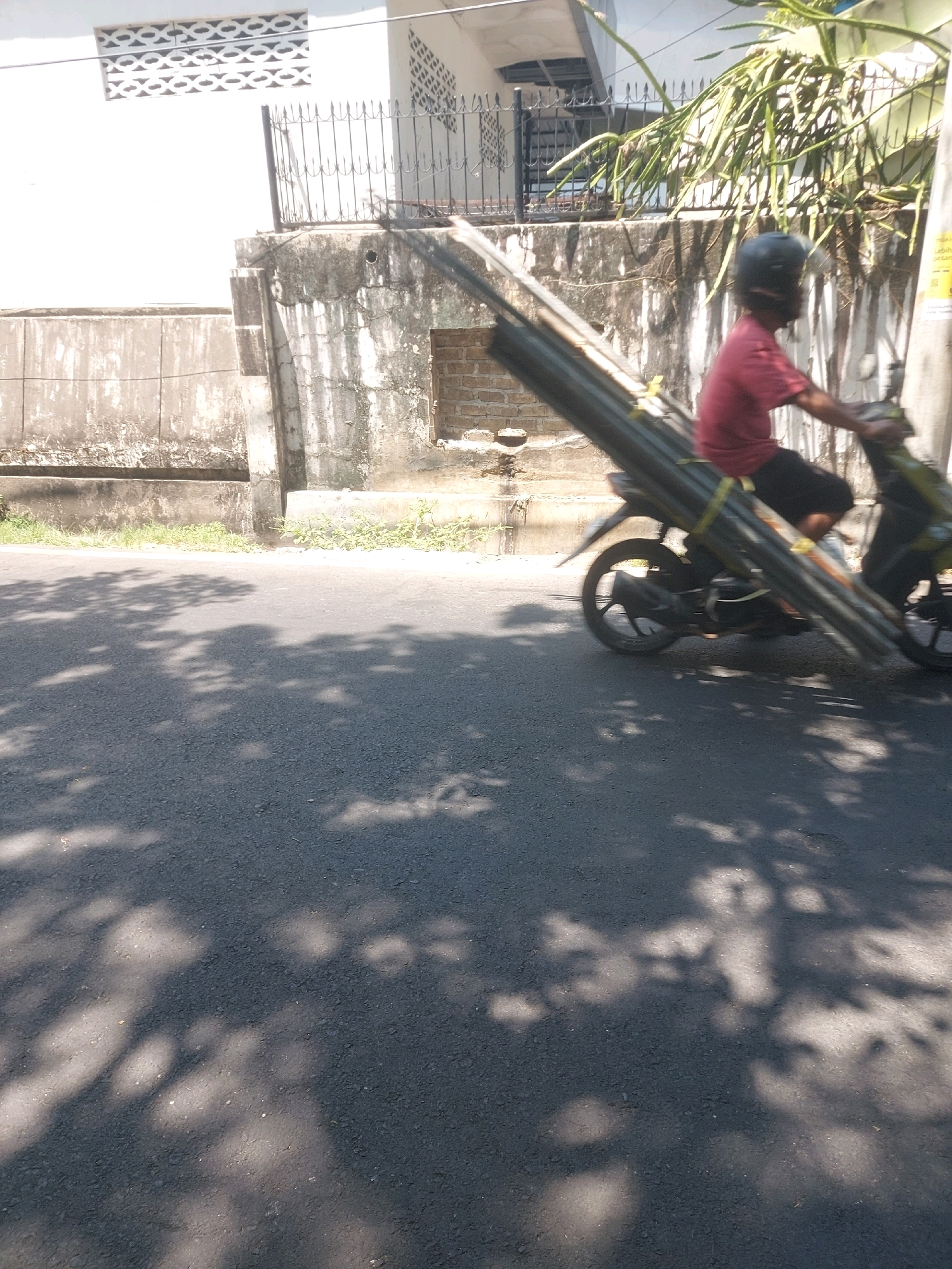 In Makassar, Indonesia, a person rides a motorcycle carrying a load of long, dark pipes. The pipes are tied to the motorcycle and extend far beyond the rear of the vehicle, creating a precarious load. The rider wears a dark helmet and a reddish-pink shirt. The motorcycle is green and black. The scene takes place on a paved road with visible shadows of trees on the asphalt. In the background, a white building with a patterned ventilation grille and a textured concrete wall suggest an urban or suburban environment. The overall impression is one of daily life and commerce in a bustling Indonesian city.