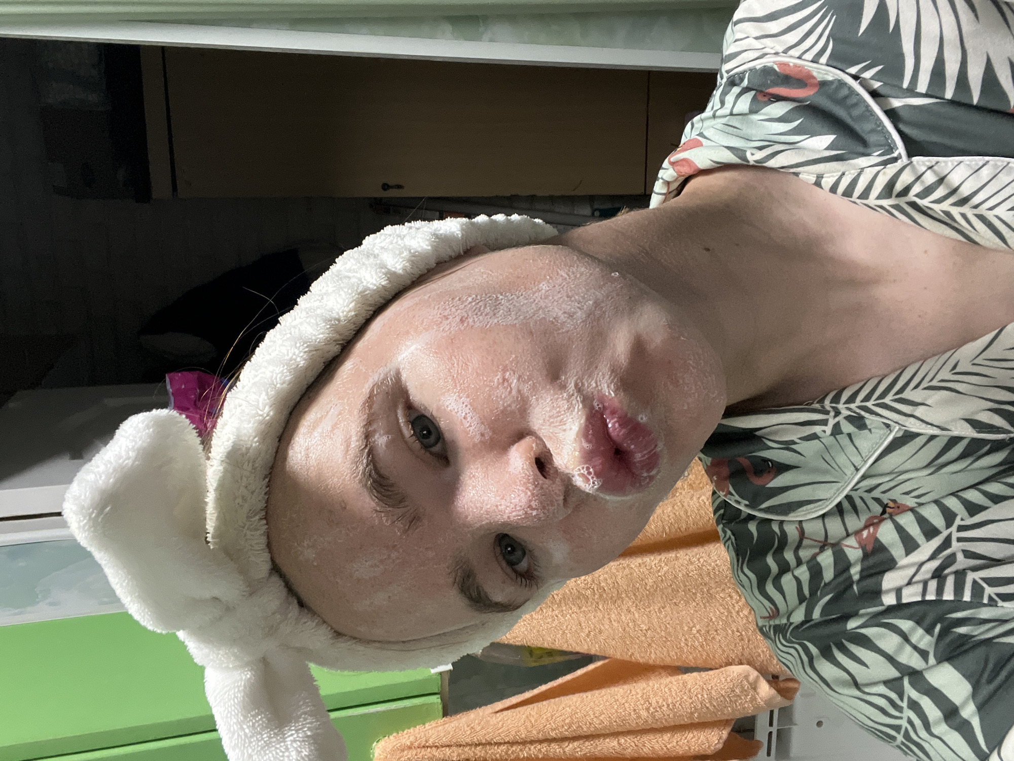 A young person with light skin and blue eyes is taking a selfie. They are wearing a white, fluffy headband with bunny ears. Their face is covered in a white facial mask, and their lips are pursed. They are wearing a pajama top with a tropical leaf and flamingo print. The background is dimly lit and appears to be a bathroom or bedroom. There are orange towels hanging on the left side of the image, and a cabinet or wardrobe in the background on the right. The image was taken in the city of Chaikovsky, Russia.