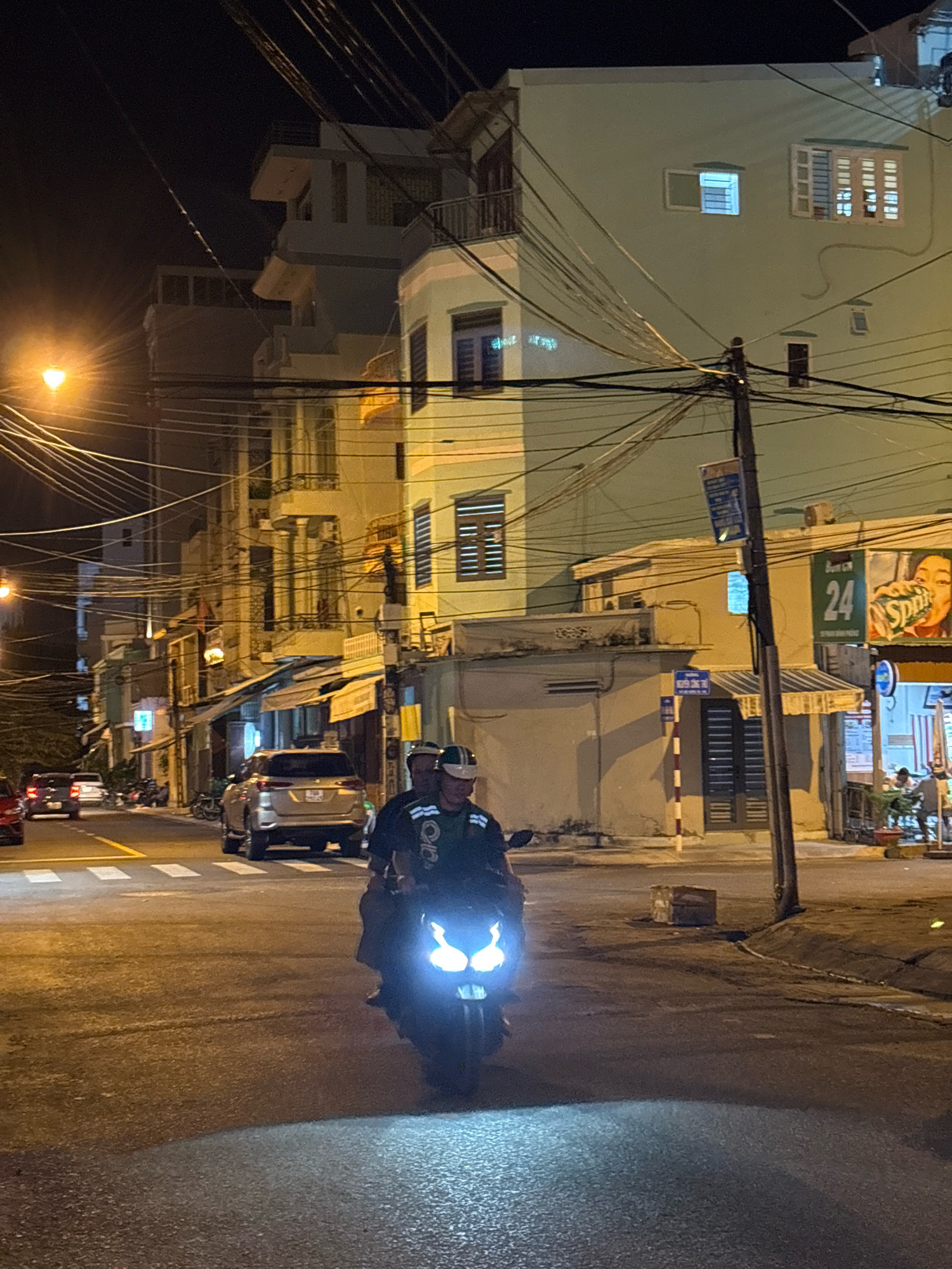 The image captures a nighttime scene on a city street, likely in Nha Trang, Vietnam, characterized by a dense network of overhead electrical wires. In the foreground, two individuals are riding a motorcycle, with their faces obscured by helmets. The motorcycle's headlights are illuminated, casting a bright glow on the asphalt road. Behind them, other vehicles, including cars and motorcycles, are visible in the middle ground and background, indicating moderate traffic. The surrounding buildings are illuminated by streetlights and interior lights, revealing their architecture, which appears to be a mix of residential and commercial establishments. The presence of awnings over storefronts and the general urban infrastructure suggest a bustling commercial district. The overall atmosphere is that of an active urban environment at night.