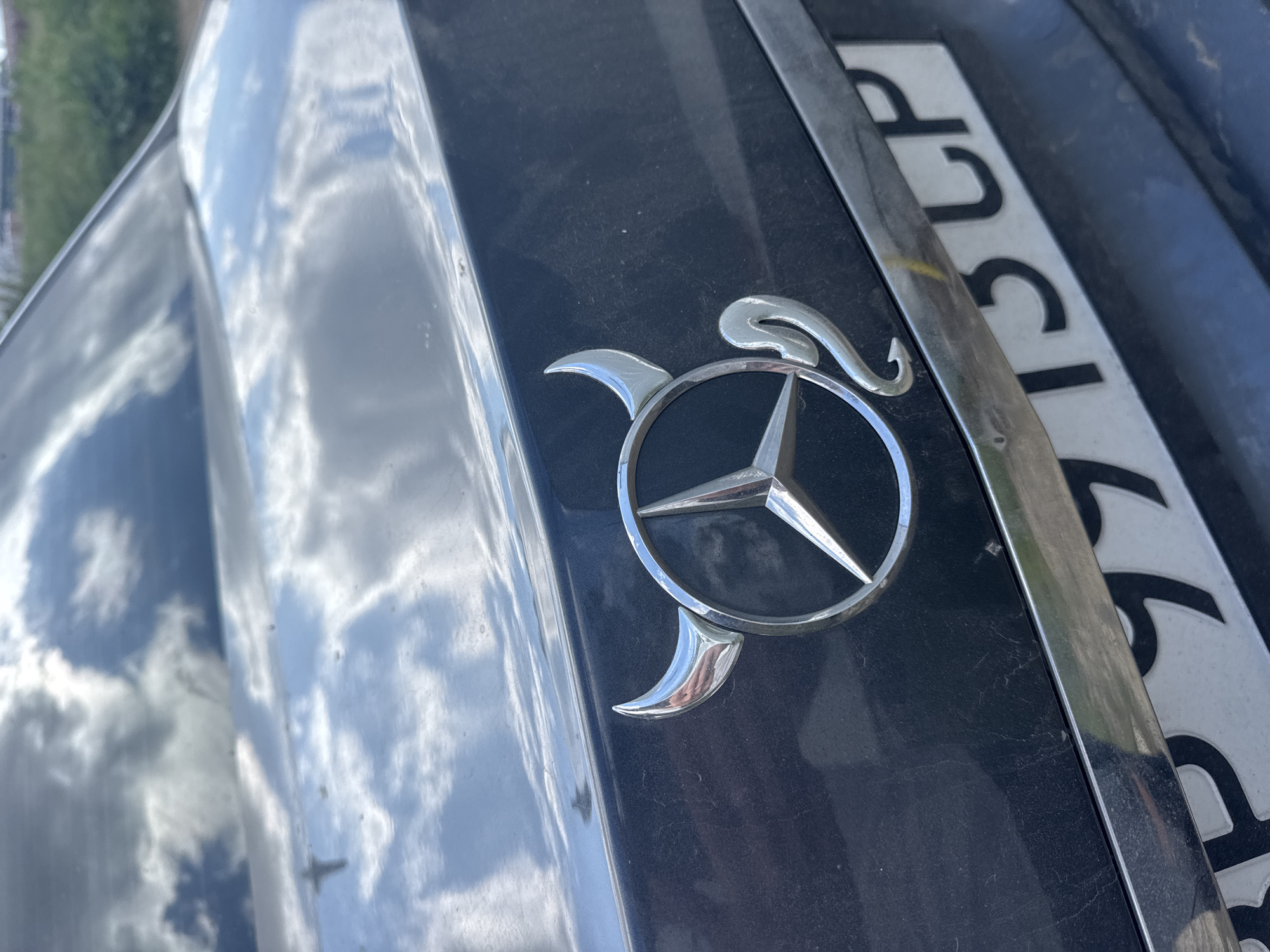 The image shows the hood of a dark-colored car, likely a Mercedes-Benz, with a modified emblem. The emblem consists of the traditional Mercedes-Benz star within a circle, but with three curved, horn-like additions attached to the circle at different points. A license plate is visible partially on the right edge of the frame. The hood of the car is dusty, and the sky is reflected on the car's surface. The location is unknown.
