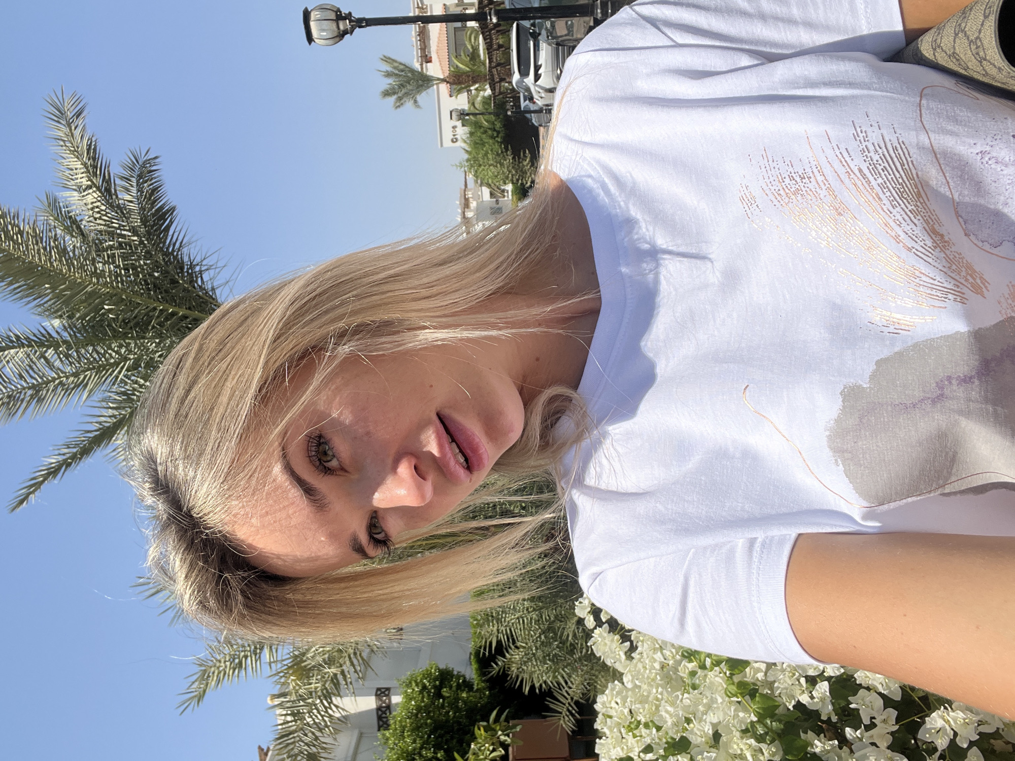 A close-up, waist-up, selfie-style photograph shows a young, fair-skinned woman with blonde hair and green eyes. She is wearing a white t-shirt with a printed design and is looking towards the camera with a slight smile. The background suggests an outdoor, sunny location in Sharm El Sheikh, Egypt, with palm trees, white buildings, and lush greenery visible. The lighting indicates a bright, daytime scene. The woman's left arm and shoulder are visible, and she appears to be holding a handbag on her right side, partially out of frame. The overall impression is one of casual leisure in a warm climate.