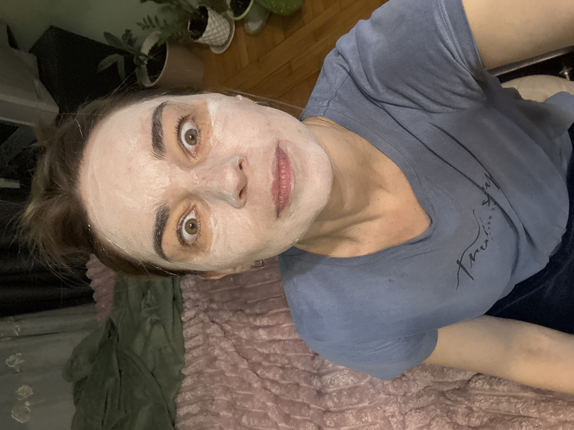A person with a white face mask is lying on a pink textured bed, wearing a blue t-shirt. A potted plant is visible in the background. The location in the city of Vsevolozhsk, Russia, is not identifiable from the image.