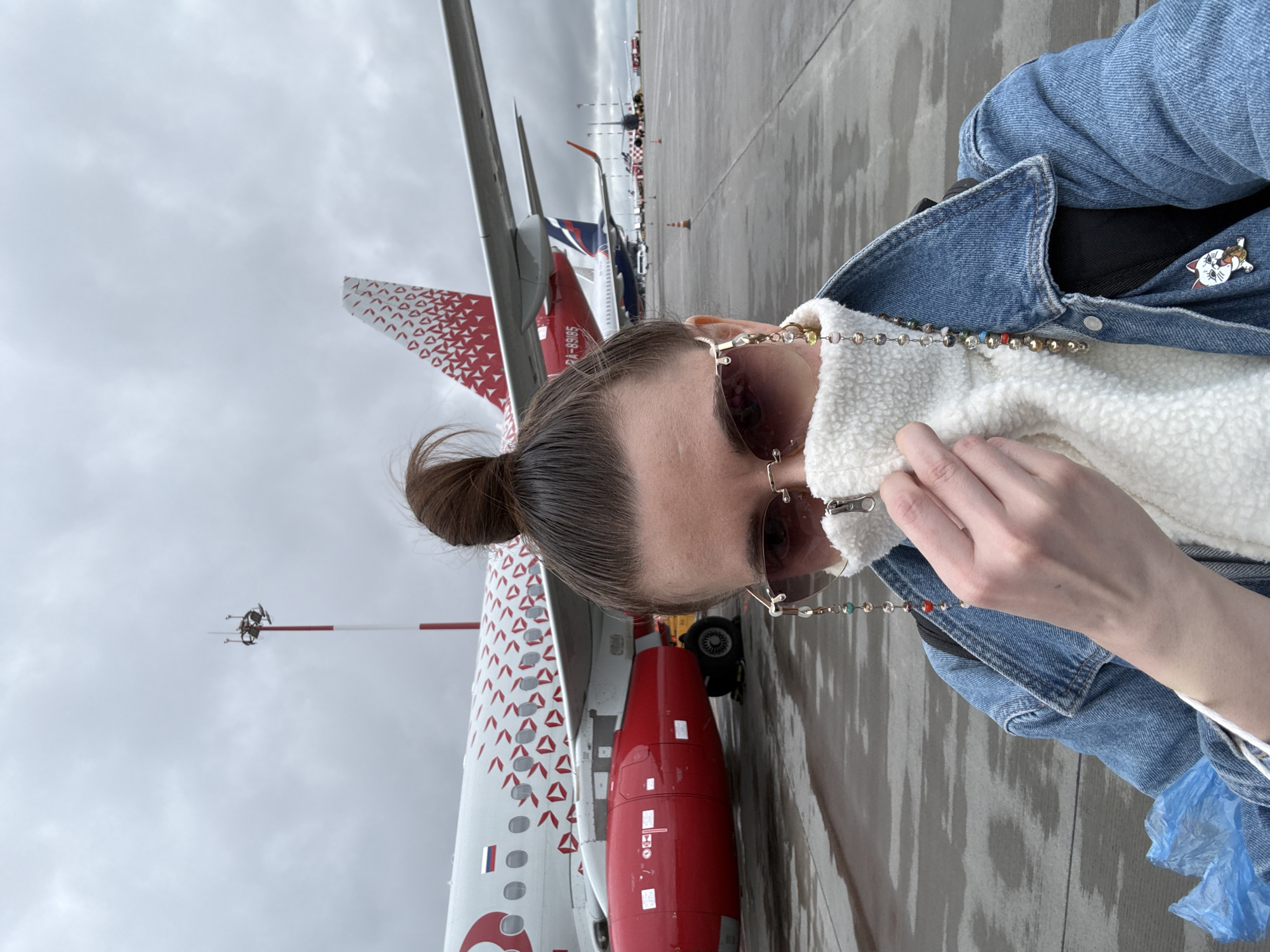A medium, eye-level shot depicts a person, presumably female, in the foreground, with several aircraft in the background. The person has brown hair styled in a bun and wears a denim jacket over a white, fuzzy top. She is also wearing sunglasses with a decorative chain. Her left hand is raised near her face, possibly adjusting her top or sunglasses. The background shows a concrete tarmac with several aircraft, the closest one being predominantly white with red markings and a red engine. The aircraft tail has a red and white pattern. In the background, other aircraft tails and orange traffic cones are visible. The sky is overcast. Based on the presence of Rossiya airlines, the location is likely at or near Sheremetyevo International Airport, located in Khimki, Russia.