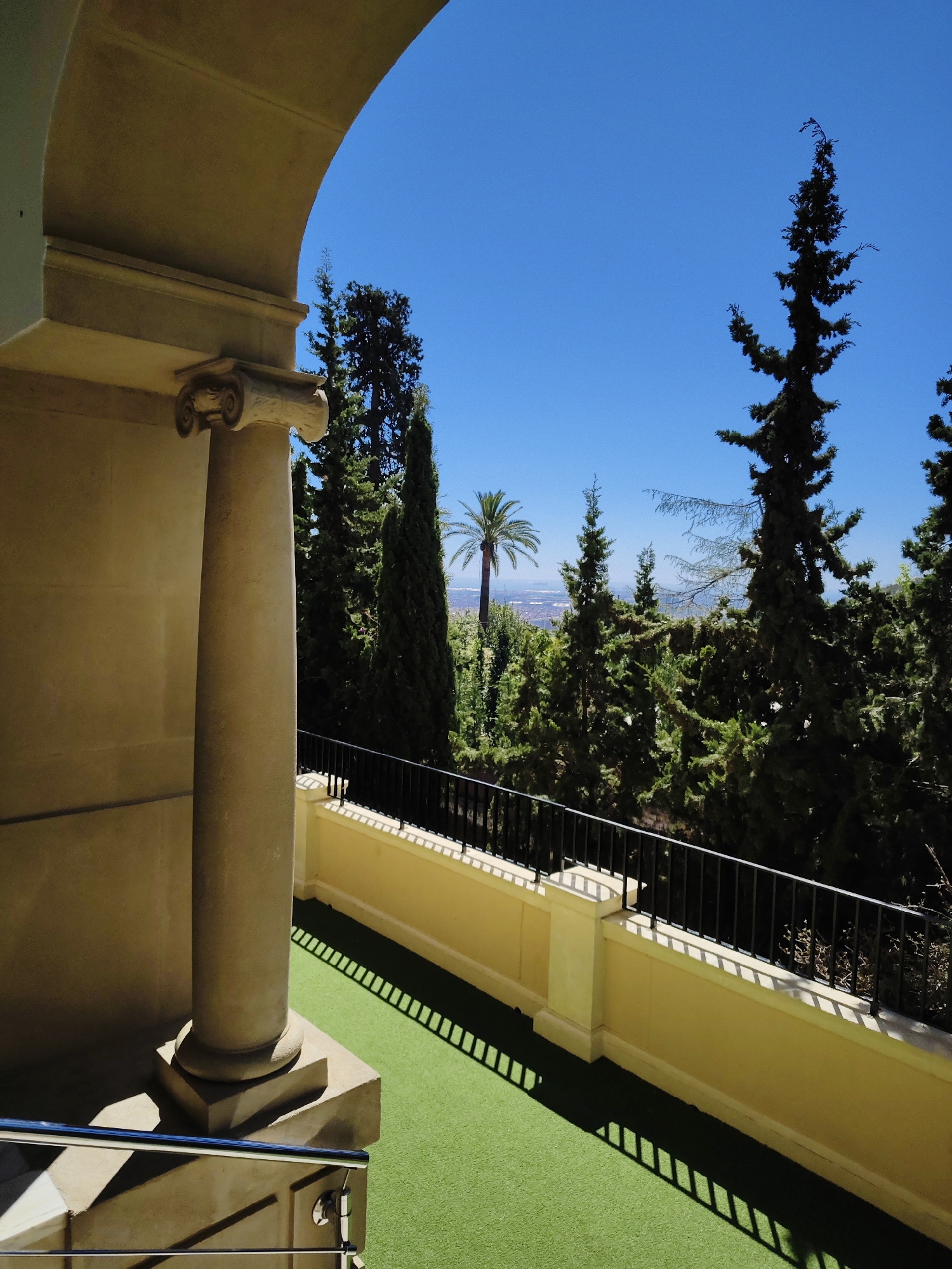 The image captures a view from a balcony or terrace, framed by an architectural archway with a classical column featuring a scroll capital. The space beyond the archway overlooks a landscape of various trees, including conifers and a palm tree, set against a clear blue sky. In the distance, a cityscape can be faintly seen, likely identifying the location as Barcelona, Spain. The balcony has a low wall with a black iron railing, casting strong shadows on the artificial green turf that covers the floor. The overall scene suggests a tranquil setting with a blend of architectural detail and natural scenery.