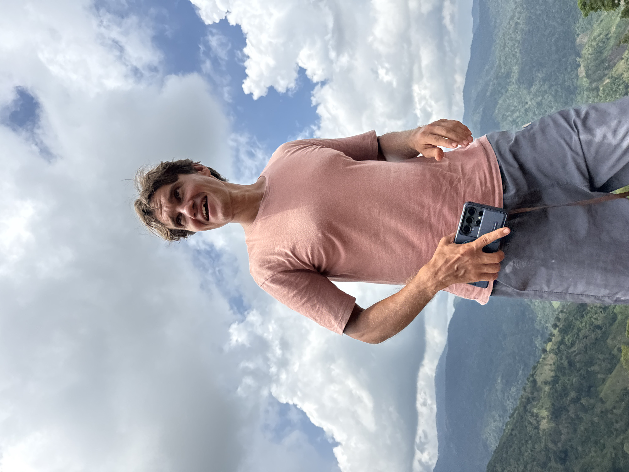 A man stands on a hill overlooking a lush, mountainous landscape in Nao Sẻ, Vietnam. He is wearing a salmon-colored t-shirt and grey shorts, and is holding a dark-colored smartphone. The sky above is filled with dramatic white clouds against a bright blue backdrop. The man appears to be looking upwards and to his left, with a slight smile on his face. The mountains in the distance are covered in dense green vegetation, with some areas appearing terraced, suggesting agricultural activity. The overall scene evokes a sense of natural beauty and peacefulness.