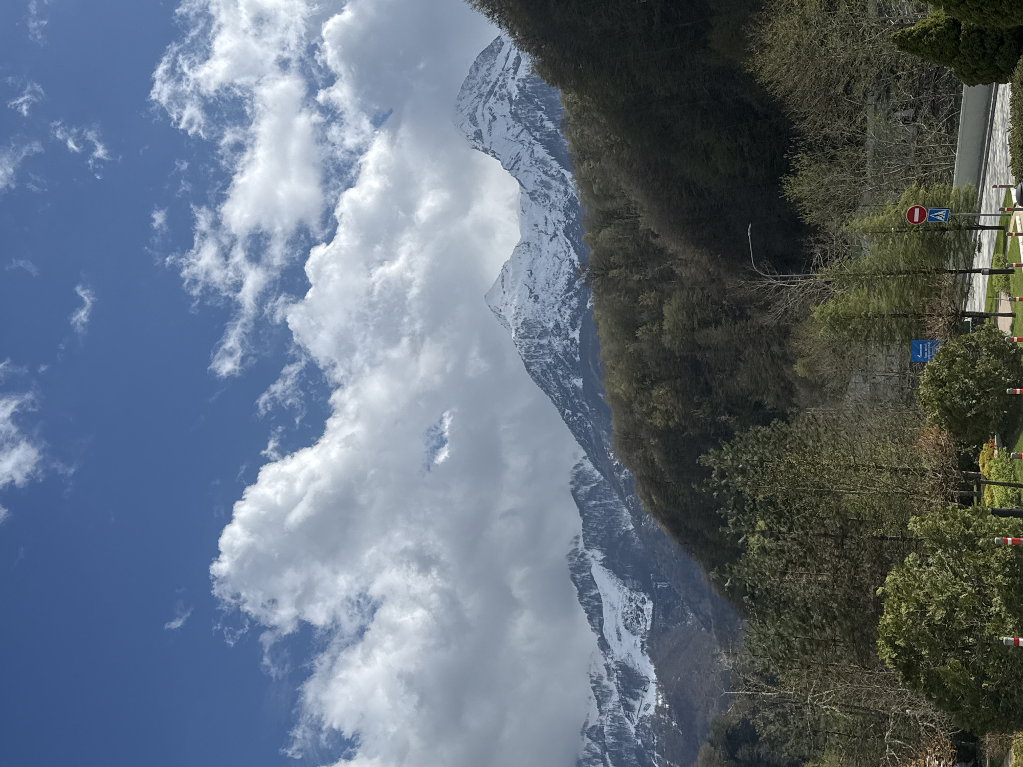 The image displays a scenic mountain landscape in Estosadok, Russia, under a bright, partly cloudy sky. Majestic snow-capped mountains dominate the central and left parts of the frame, their peaks partially obscured by large, fluffy white cumulus clouds. The sky above is a clear blue, with scattered wisps of cirrus clouds.
To the right, a densely forested slope with both green coniferous trees and bare deciduous trees descends towards the foreground, suggesting a spring season. In the lower right, a man-made area is visible, featuring a paved road or pathway. Along this path, there are trimmed bushes and trees, a street lamp, and several road signs. A prominent red circular sign with a white horizontal bar indicates "No Entry" or "Do Not Enter." Below it, a blue rectangular sign with an indistinguishable white symbol or text is present. Red and white striped barrier poles line the edge of the paved area.
The time of day appears to be late morning or early afternoon, indicated by the bright, overhead illumination. The scene conveys a sense of natural grandeur and tranquility, with no people visible.