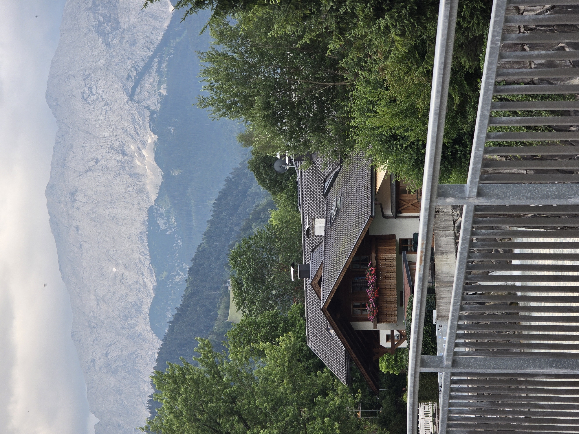 The image depicts a view of a multi-story house with a dark tiled roof, a wooden balcony adorned with flower boxes filled with pink flowers, and wooden shutters on the windows. The house is nestled amongst lush green trees and is situated in front of a majestic, grey, rocky mountain range under a cloudy sky. In the foreground, a metal railing and vertical bars of a bridge or walkway are visible, overlooking a body of water. The scene is likely located in or near Garmisch-Partenkirchen, Germany, given the characteristic alpine landscape.