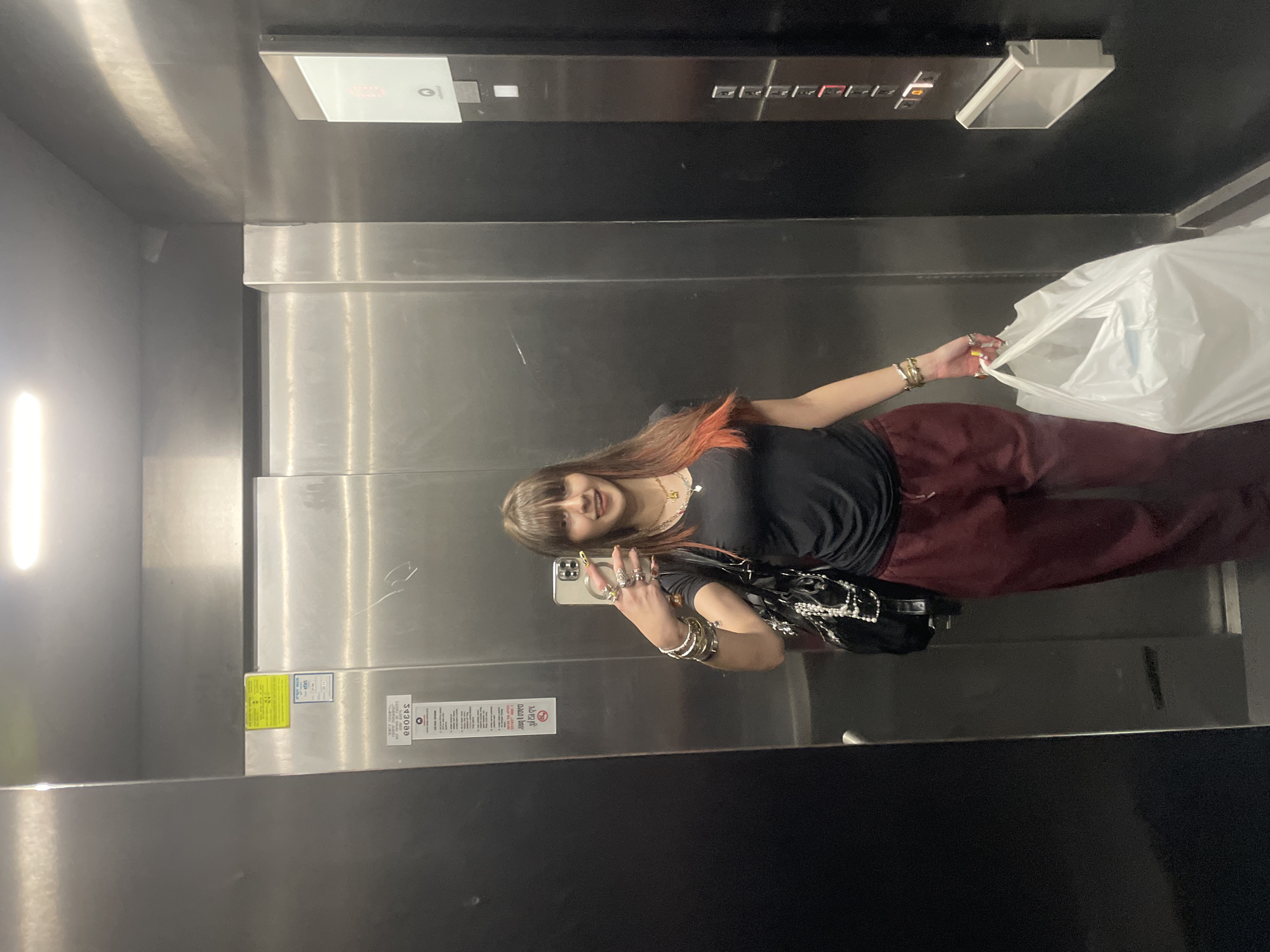 A young woman is taking a selfie in an elevator. She is holding a white plastic bag in her left hand. The elevator has stainless steel walls and a control panel with floor numbers. There is no indication of the elevator's location in Raanana, Israel, so this cannot be confirmed.