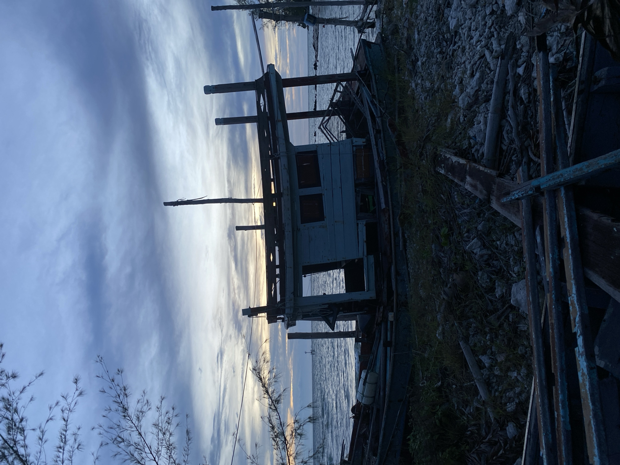 An abandoned, weathered wooden fishing boat rests partially on a rocky shore with tall, thin grass. The boat is painted light blue and appears to be in disrepair, with a broken canopy and exposed wooden beams. In the background, the ocean stretches to the horizon under a cloudy, twilight sky. A small lighthouse or buoy is visible in the distance on the water. Debris, including broken pieces of wood and vegetation, litters the ground around the boat. The scene conveys a sense of desolation and the passage of time.