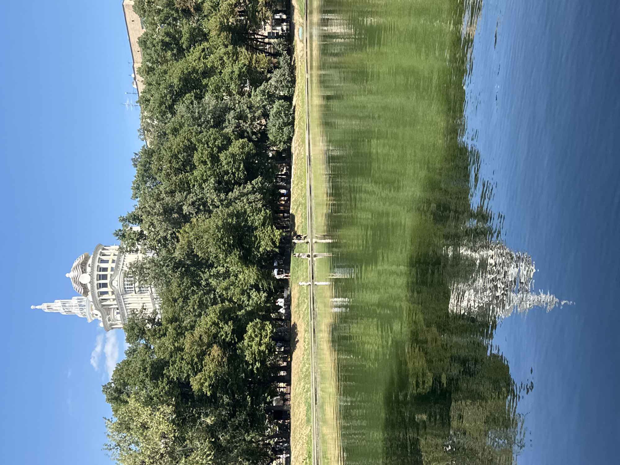 The image depicts a wide, still body of water, likely a pond or lake, with a clear blue sky overhead. The water's surface reflects the surrounding environment, including a dense line of lush green trees that form a natural barrier along the far bank. Behind the trees, a prominent, ornate white building with a distinctive spire and domed roof stands tall. This building is a landmark, the "House of the Architect" or "Dom Arkhitektora," located in Moscow, Russia. Several small groups of people are visible on the grassy area between the trees and the water, appearing to be relaxing or strolling. A few individuals are also seen walking along the edge of the water. The scene conveys a peaceful, urban park atmosphere on a sunny day.