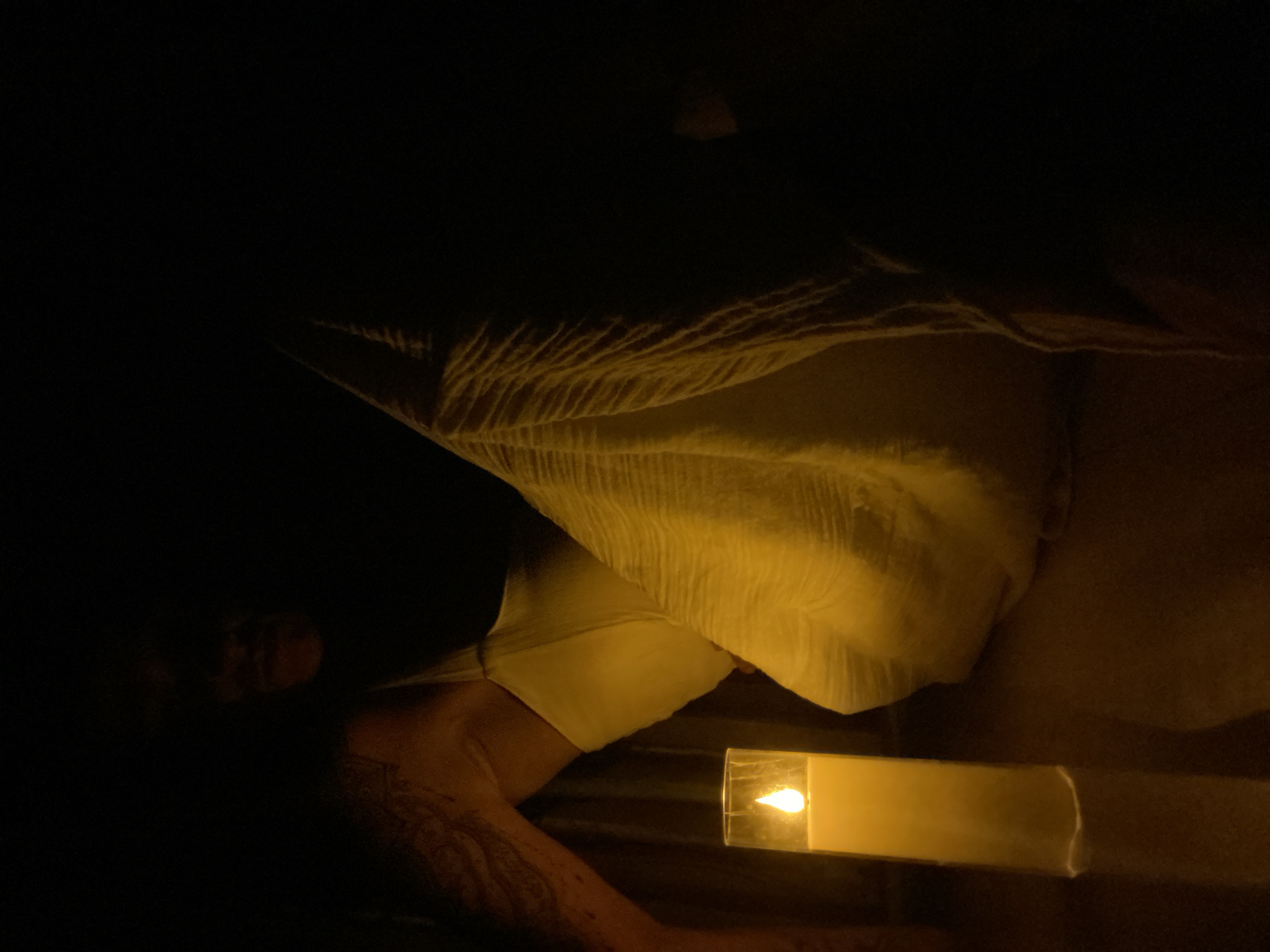 The image shows a person sitting in a dimly lit room, possibly a bedroom or a quiet space, with a single lit candle in the foreground. The candle is in a glass container, casting a warm, yellow glow on the surroundings. The person is partially visible, with their face obscured by shadow, but their arm and shoulder are illuminated by the candlelight. They appear to be wearing a white garment. The overall mood is serene and introspective, with the darkness of the room emphasizing the light of the candle. The image does not provide any specific details or visual cues to indicate a location in Mũi Né, Vietnam.