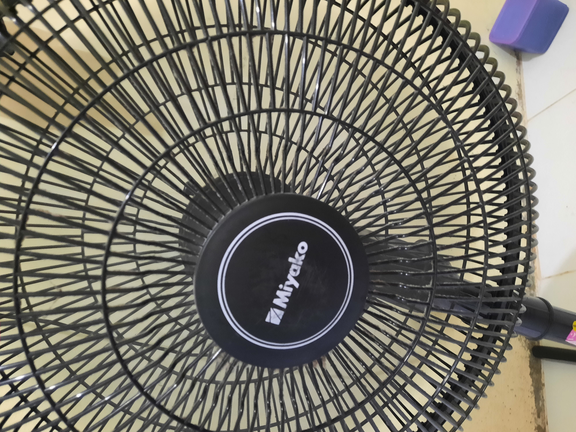 The image displays a close-up view of a black oscillating fan, specifically focusing on its front grille and central casing. The fan features a circular logo with the brand name "Miyako" written in white text, accompanied by a triangular emblem. The fan's grille is composed of numerous thin, parallel metal bars arranged in a radial pattern. The background appears to be a wall or surface with a light yellow hue. The fan is positioned in an indoor setting. No people are visible in the image, and no actions or interactions are depicted. The overall scene is static and observational. The location of Lhokseumawe, Indonesia, is not visually represented in this image, as there are no identifiable landmarks or contextual clues to suggest this specific geographical area.
