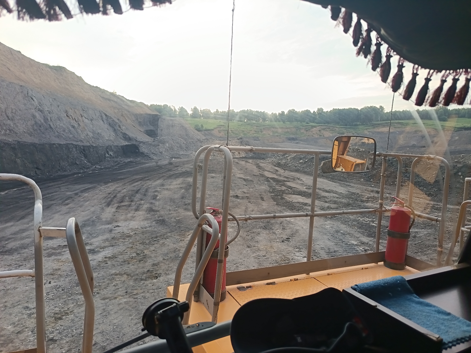 The image, taken from inside a vehicle cabin, shows a view of a large open-pit mine. The foreground features part of the vehicle's interior, including a steering wheel mechanism, a blue cloth, and a red fire extinguisher mounted on a railing. Beyond the vehicle's window, a vast expanse of the mine is visible. The terrain is characterized by steep, excavated slopes covered in dark gray soil and rocks, with tire tracks evident on the ground. In the distance, a line of trees and green vegetation creates a stark contrast with the barrenness of the mine. The sky is bright and overcast, suggesting daylight. There are no people visible in the image. The location is identified as Kyrgai, Russia.