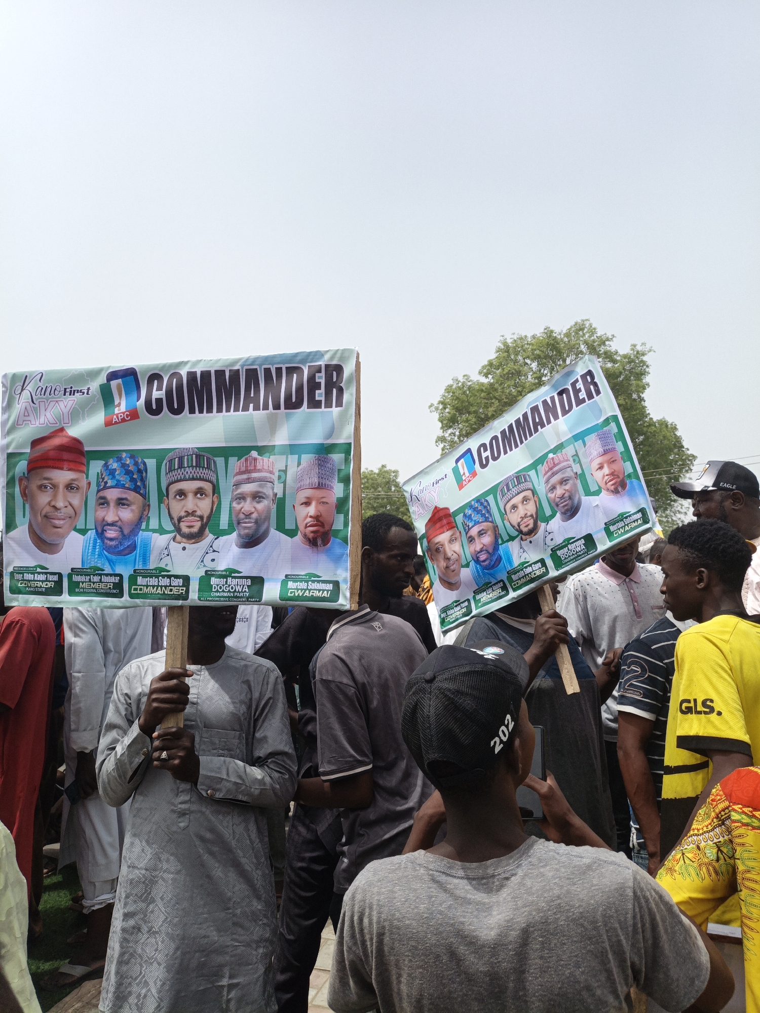 A medium shot captures a crowd of people in Tarauni, Nigeria, during a political event. Two individuals hold signs. Each sign features a banner with the words "Kano First," "APC," and "COMMANDER" in bold letters. Beneath the banners, the signs display photographs of five men identified as Engr. Abba Kabir Yusuf (Governor), Abubakar Kabir Abubakar (Member), Murtala Sule Garo (Commander), Umar Haruna Dogowa (Chairman Party), and Murtala Sulaiman Gwarmai. The man on the left is wearing a grey colored robe. The man on the right is taking a picture of the sign. A black baseball cap with "202" is displayed on the back of the man's head. A sign with "EAGLE" is shown on the right side of the image. A yellow shirt with "GLS." is also seen on the right side of the image. The event occurs outdoors under a clear, bright sky.