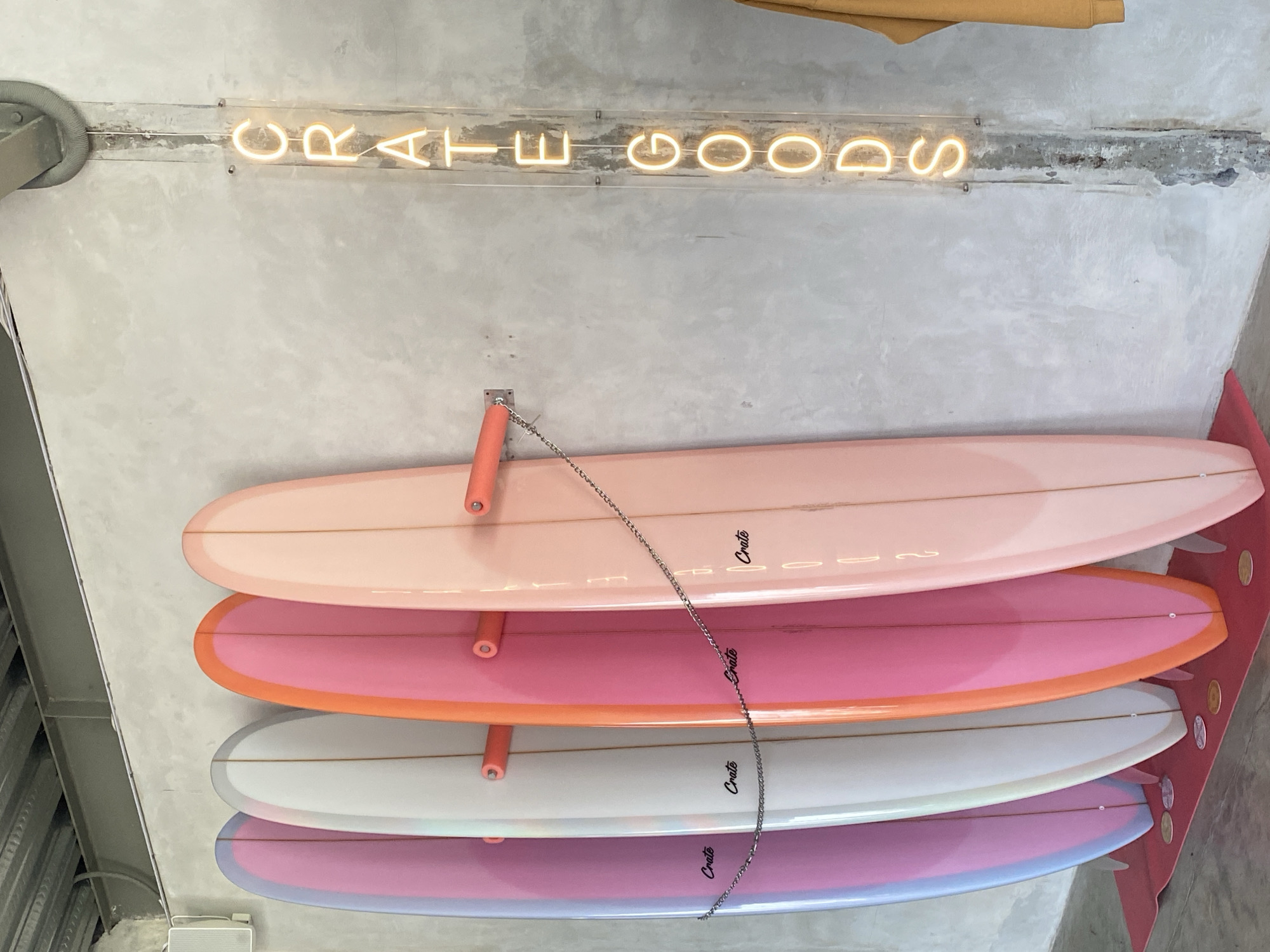 The image shows a stack of surfboards displayed on a wall. The surfboards are in varying shades of pink, white, and lavender. The surfboards are held in place by peach-colored arms. The sign "Crate Goods" in neon lighting is visible above the surfboards. A metal chain runs vertically along the surfboards. The location is Mengwi, Indonesia.