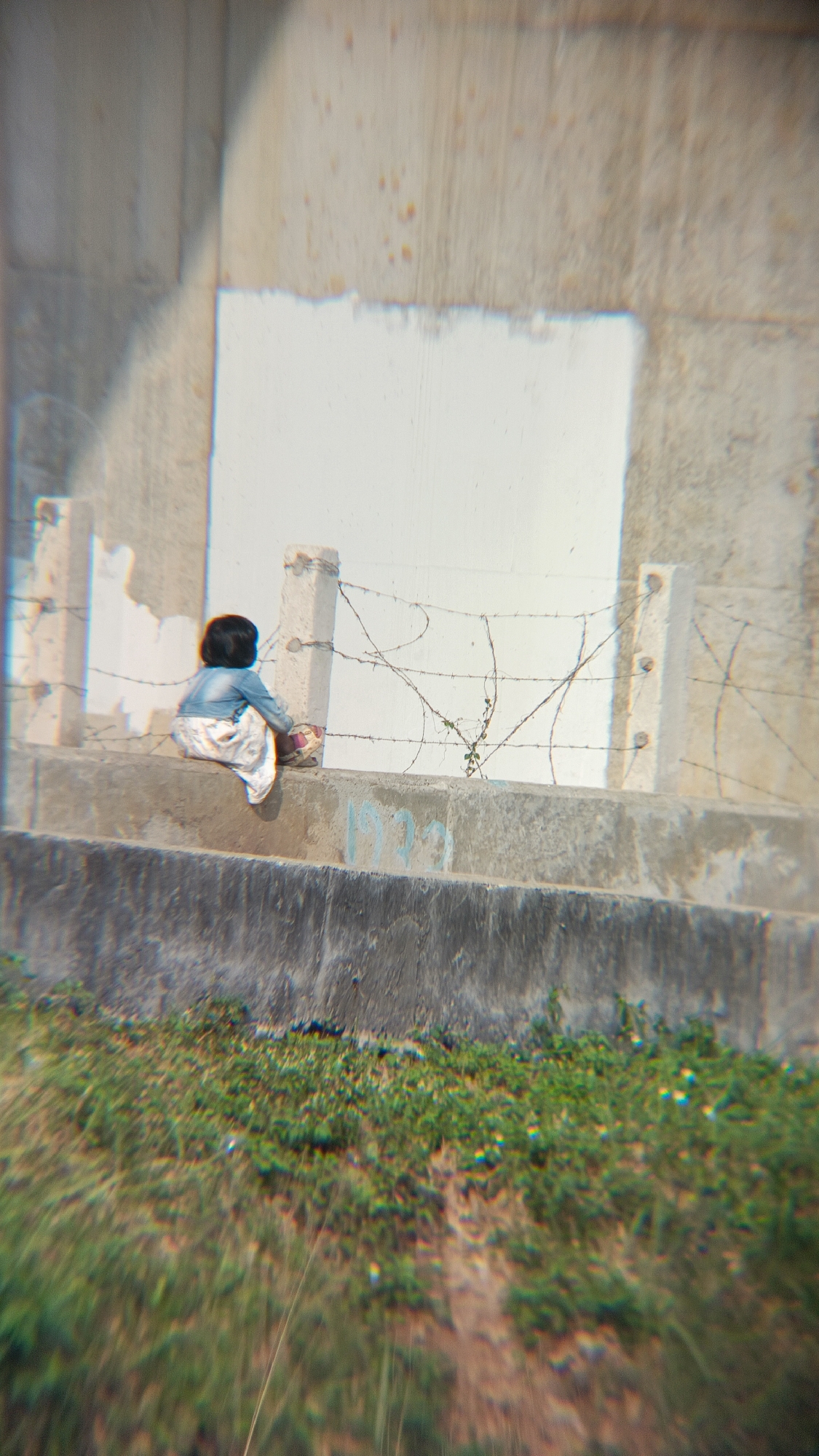 A solitary young child with dark hair, wearing a denim jacket and a patterned white skirt, sits on a concrete ledge. They are facing away from the camera, towards a fence constructed with barbed wire and supported by concrete posts. Behind the fence, a large, light-colored, rectangular area is painted on a concrete wall. In the foreground, out-of-focus green grass and brown earth are visible. The scene appears to be outdoors, possibly in an urban or semi-urban setting within Margajaya, Indonesia, given the concrete structures and the presence of barbed wire, which might suggest a restricted or demarcated area. The lighting suggests it is daytime.
