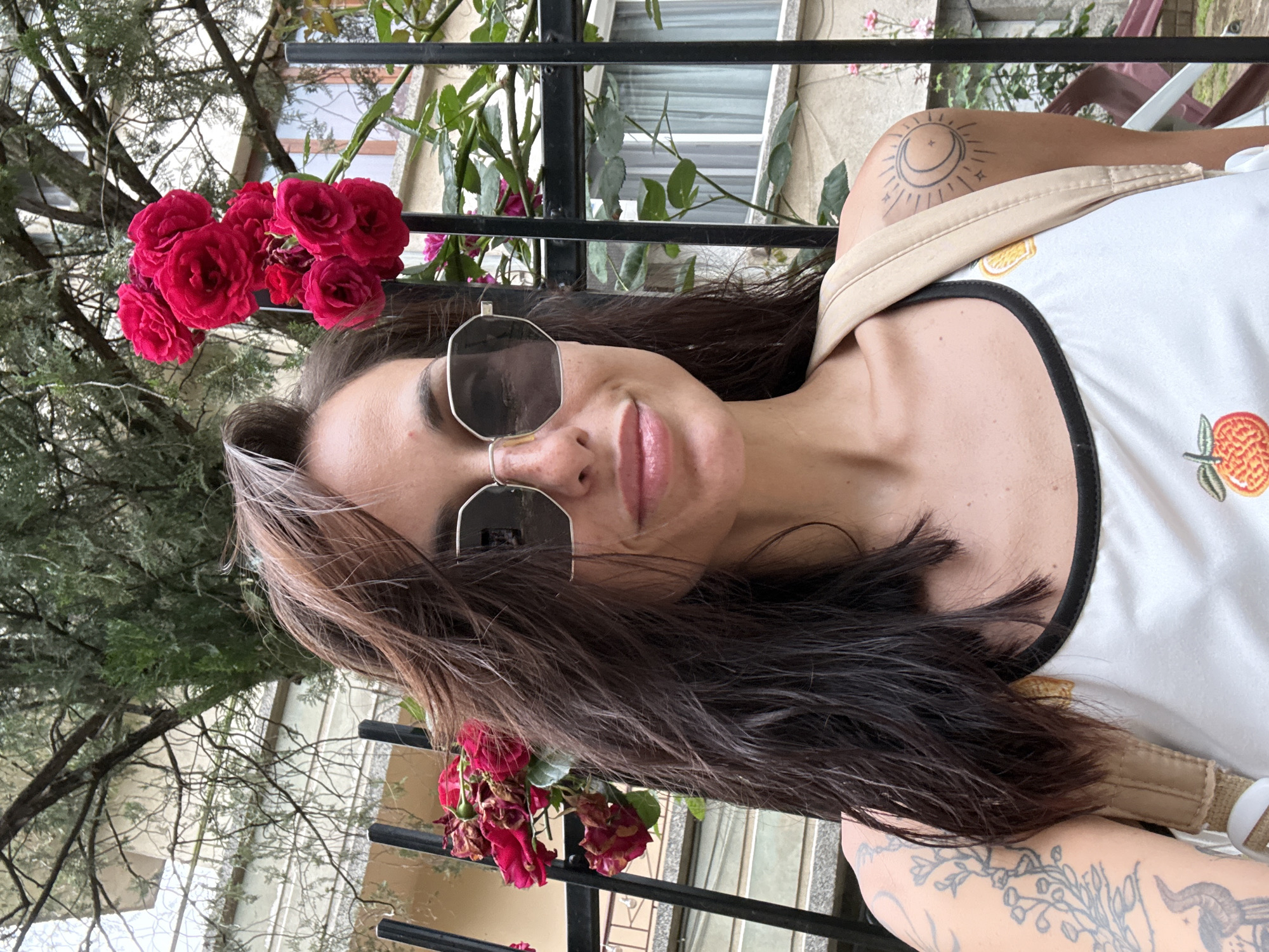A female subject, appearing to be in her 20s or 30s, is depicted in a medium shot, looking directly at the camera with a subtle smile in the city of Pleven, Bulgaria. She has dark, shoulder-length hair, possibly wet, with lighter strands at the crown, and wears octagonal dark-lensed sunglasses with light frames. Her attire consists of a white tank top with black trim and a fruit motif. Tattoos are visible on both arms: a botanical and bird design on the left, and a sun-and-crescent moon motif on the right shoulder, over which a light beige backpack strap is positioned. The subject is situated outdoors behind a black metal fence. Vibrant red roses and green foliage are prominent to her right, while wilted red roses are visible to her left. The background reveals a light-colored building with windows and trees, all under an overcast sky.